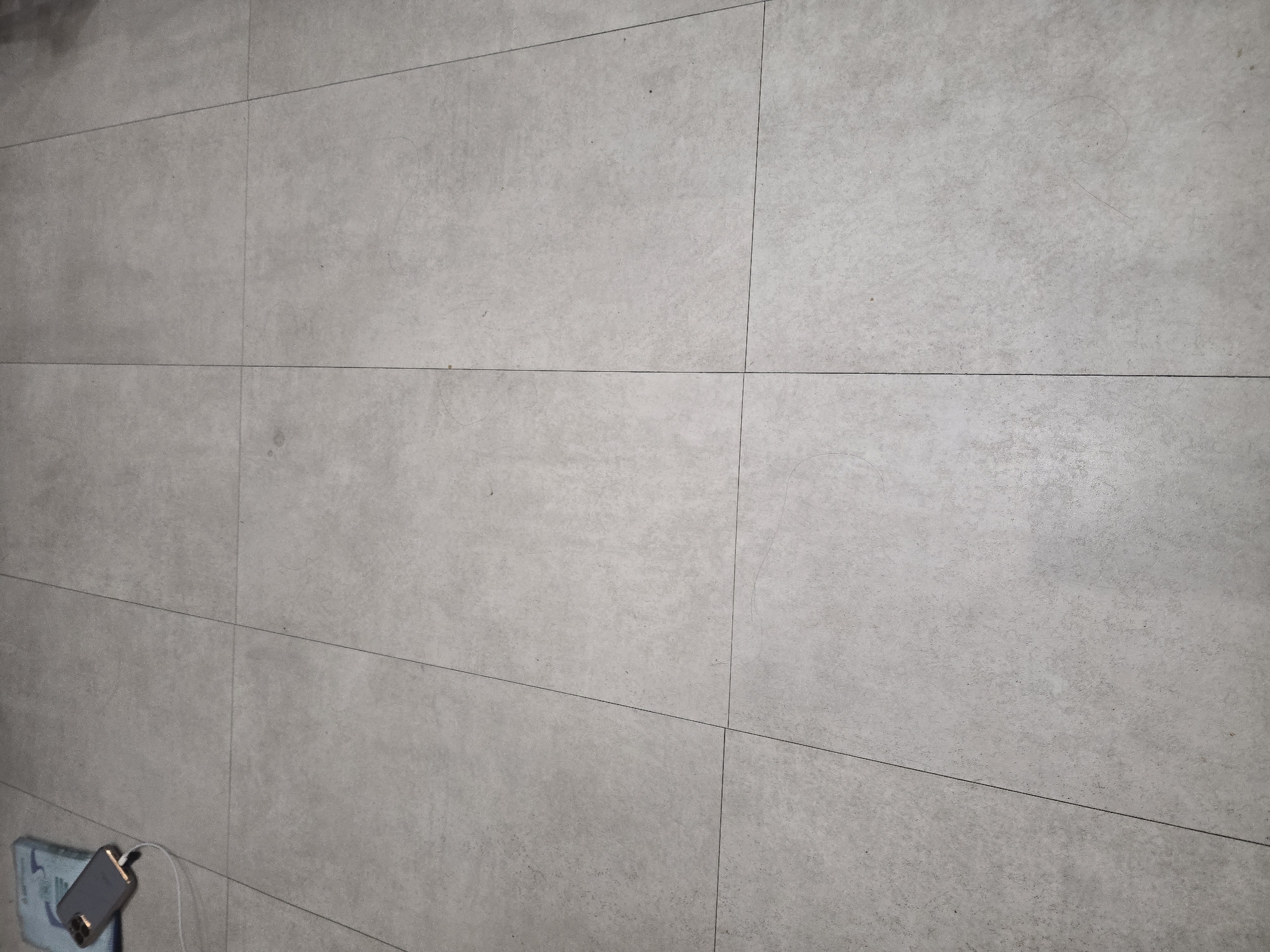 An overhead shot shows a grey tiled floor. In the upper left corner, a silver smartphone is plugged into a blue case with a white charging cable. The phone's screen is dark. The tiles are large and square, laid out in a grid pattern with thin grout lines. The lighting suggests an indoor environment, possibly a home or office. There is no indication of the specific location in Ulan-Ude, Russia, beyond the tiled floor. The scene is static and shows no human presence or activity.