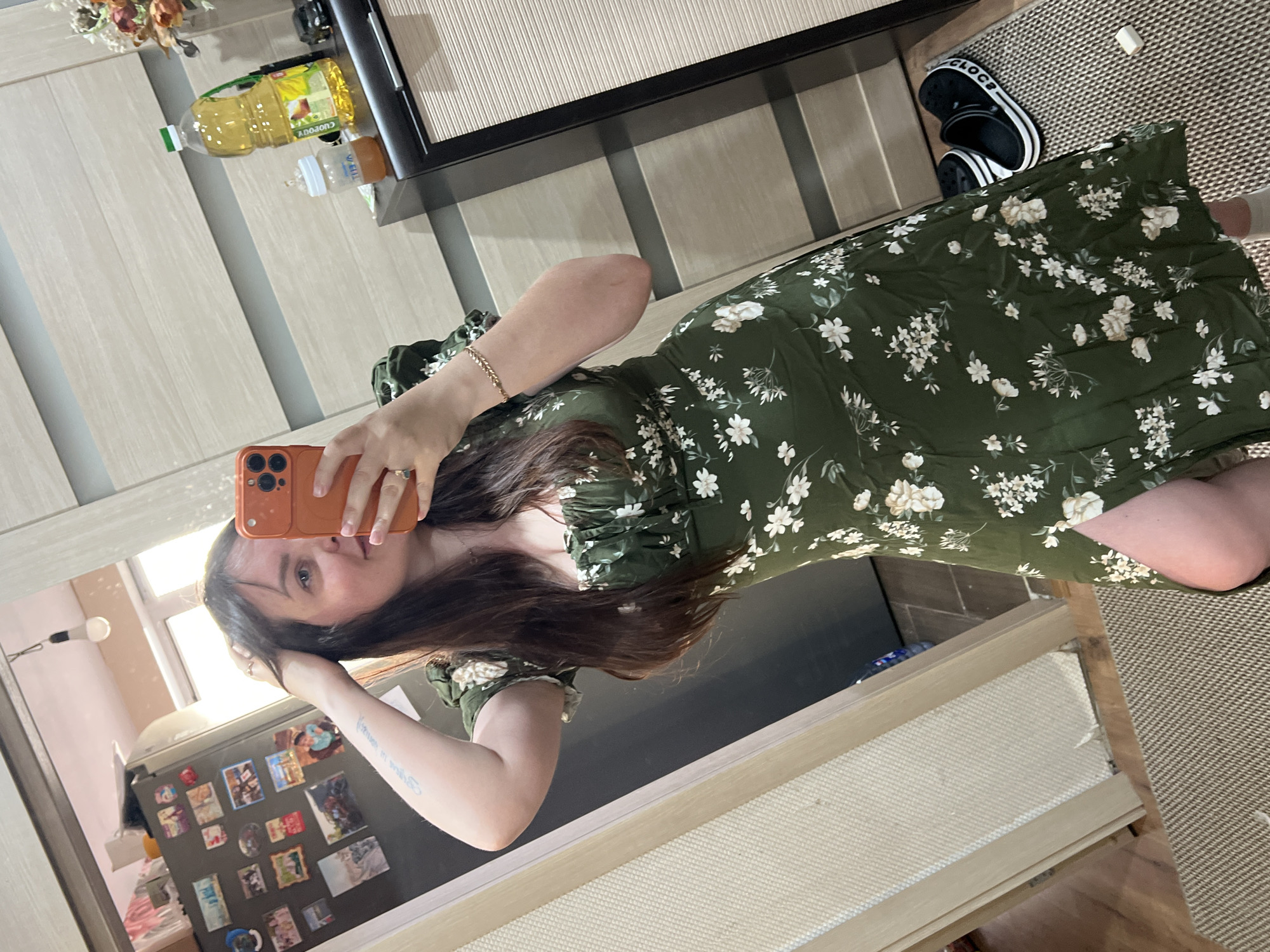The image is a vertically oriented selfie taken in a room, likely a home. The primary subject is a young woman with long, dark hair, wearing a green floral-print dress. She is holding an orange smartphone to take the picture, and her reflection is visible in a large mirror in front of her.
The setting appears to be a living area. Behind the woman, we see a wooden cabinet with a striped blind, a bottle of yellow liquid (possibly cooking oil), and a baby bottle. To the right of the woman, a pair of black Crocs shoes with white stripes are visible on a woven rug. Further in the background, a refrigerator is covered with numerous magnets and photographs. The overall lighting suggests daytime, and there are no clear indications of the weather. The woman's pose, with her hand touching her hair, and her facial expression are neutral, suggesting a casual or everyday moment. The visible text includes "CROCS" on the shoes and "CUDENGO" on the bottle of oil, though the latter is partially obscured. The location context is Noviy, Russia.