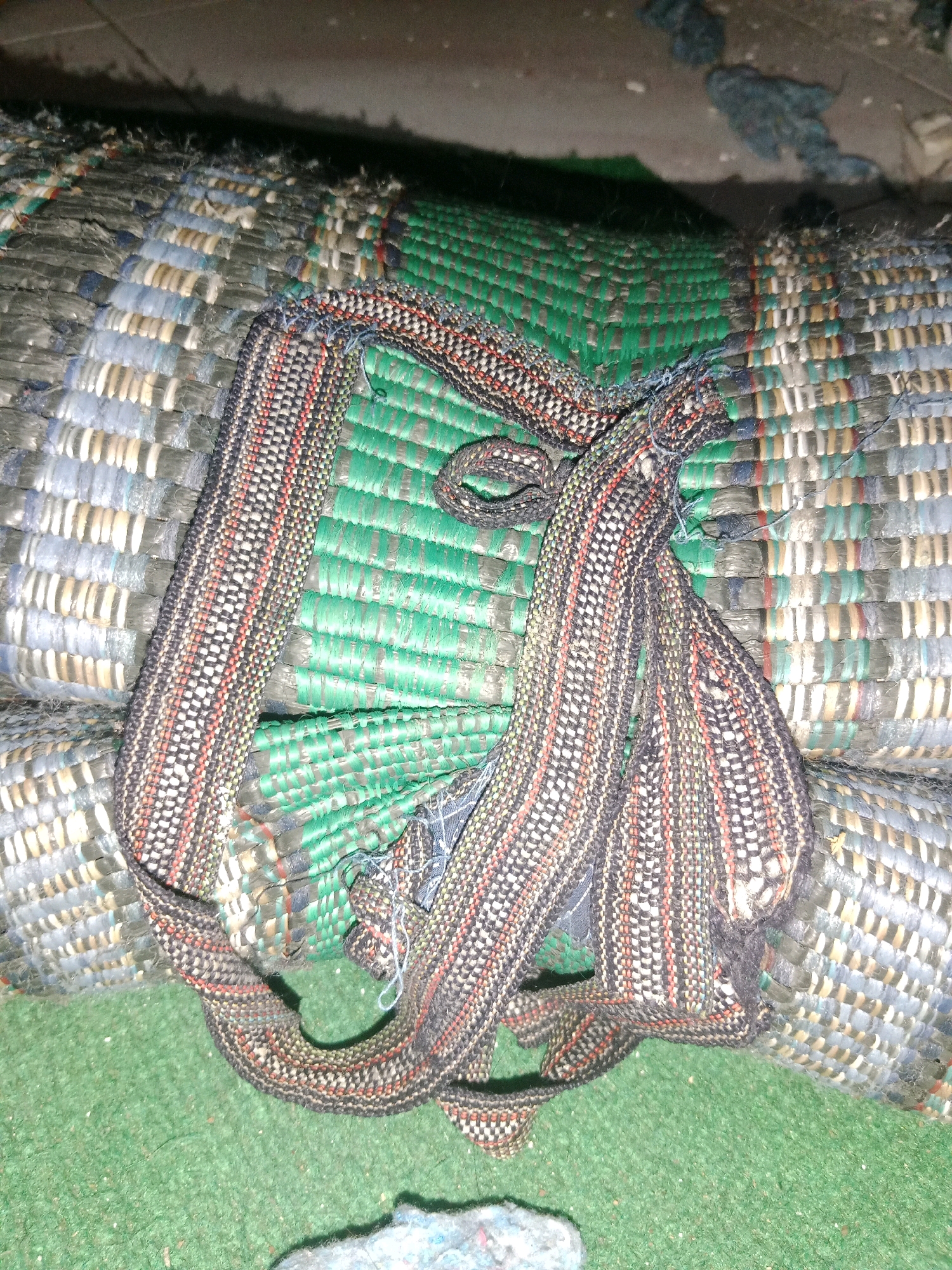 A woven mat, possibly for sleeping, is rolled up and secured with a strip of fabric. The mat displays a variety of colors and textures, with bands of green, gray, blue, and white woven together. The securing strip is dark with red and white vertical stripes. The mat appears to be made of plastic or similar synthetic material. The scene is set on a green carpeted surface, with the background showing a darker, possibly concrete or dirt, area. The lighting is dim, suggesting an indoor or evening setting. The image is taken from a close-up perspective, focusing on the details of the mat's texture and construction. The location is identified as Ringinrejo, Indonesia.