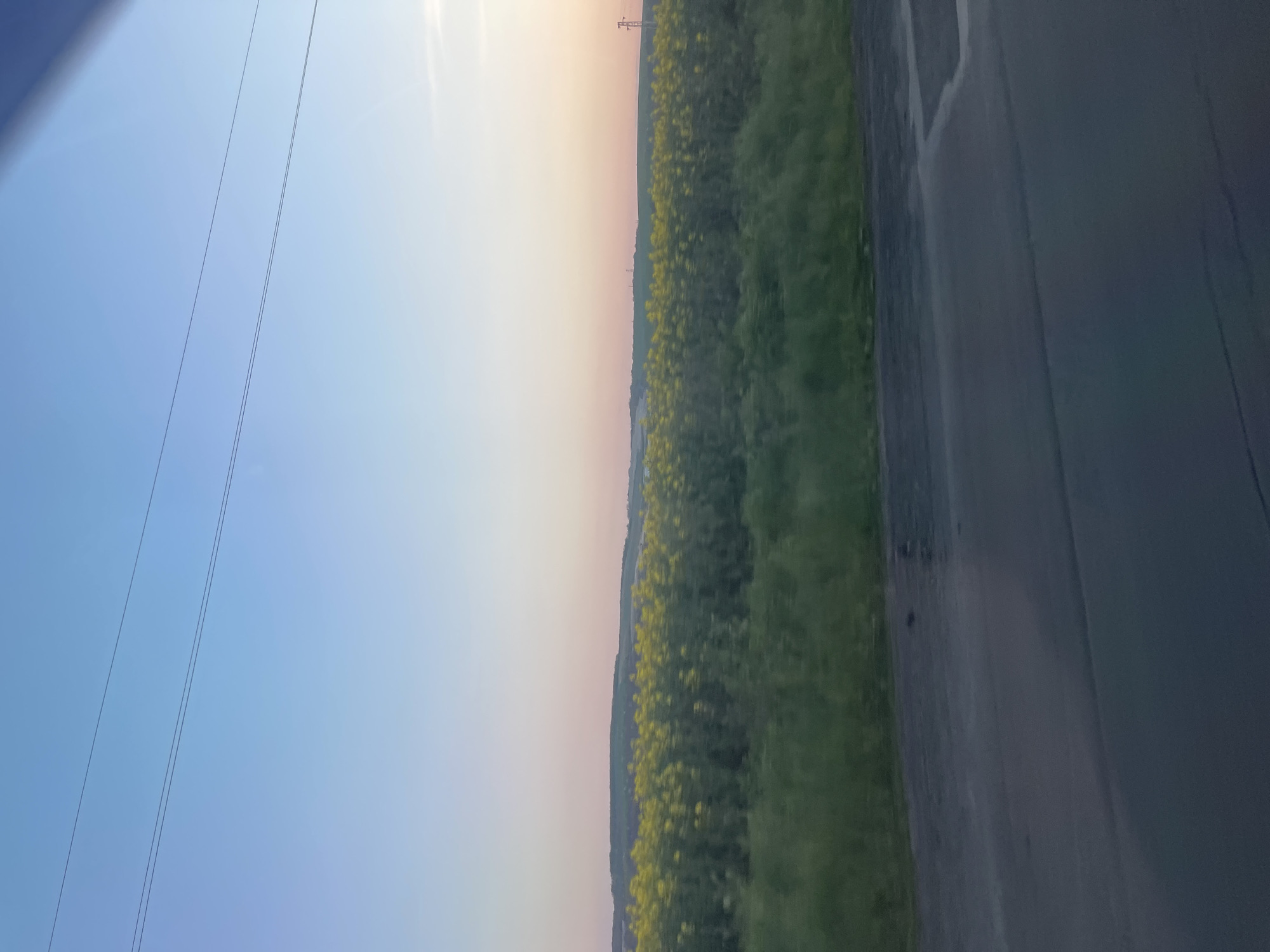 The image shows a landscape. On the left is a blue sky. A field of green vegetation with yellow flowers is adjacent to the sky. A gray asphalt road is to the right of the field. Power lines are visible against the sky. A power transmission tower can be seen in the distance. The location is assumed to be near Bratovo, Bulgaria, based on the user's prompt.