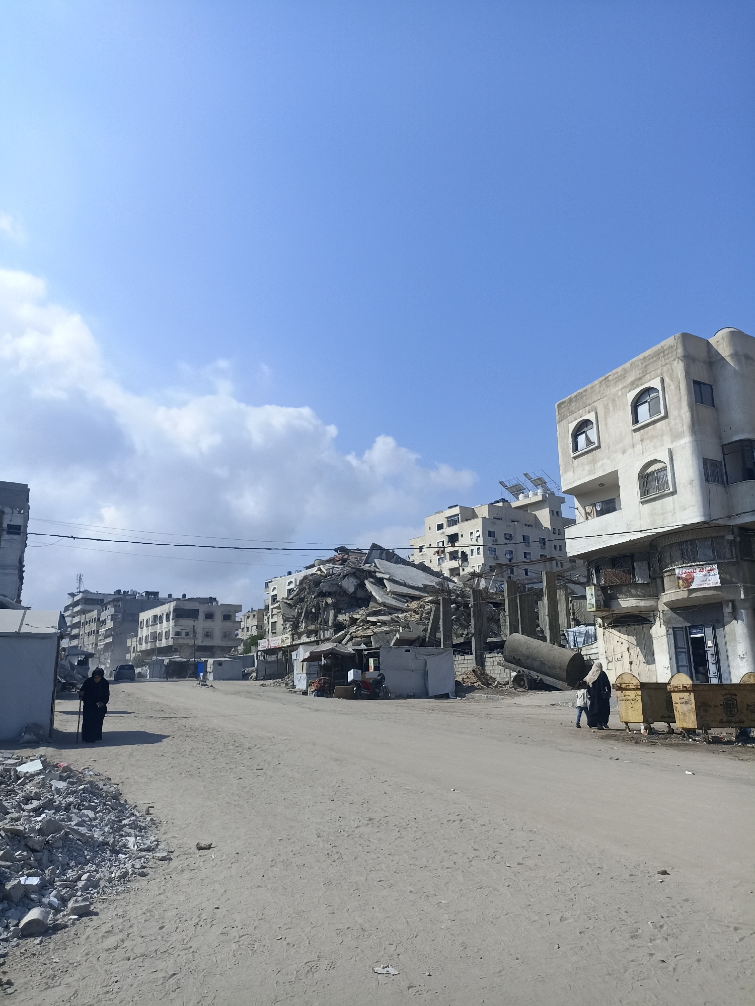 The image depicts a street scene in Gaza City, Palestinian Territories, characterized by significant destruction and debris. In the foreground, a dirt road is lined with piles of rubble on the left and extends into the distance. Several buildings are visible, many of which appear damaged or destroyed, with collapsed sections and exposed structures. One building on the right is largely intact and has shops on its ground floor. Two women, dressed in dark clothing and headscarves, are present in the scene. One woman stands near a pile of rubble on the left side of the road, while another is seen further down the road, accompanied by a child. Yellow industrial bins are situated near the buildings on the right. The sky is blue with scattered clouds. The overall environment suggests a post-conflict or disaster-affected area within an urban setting.
