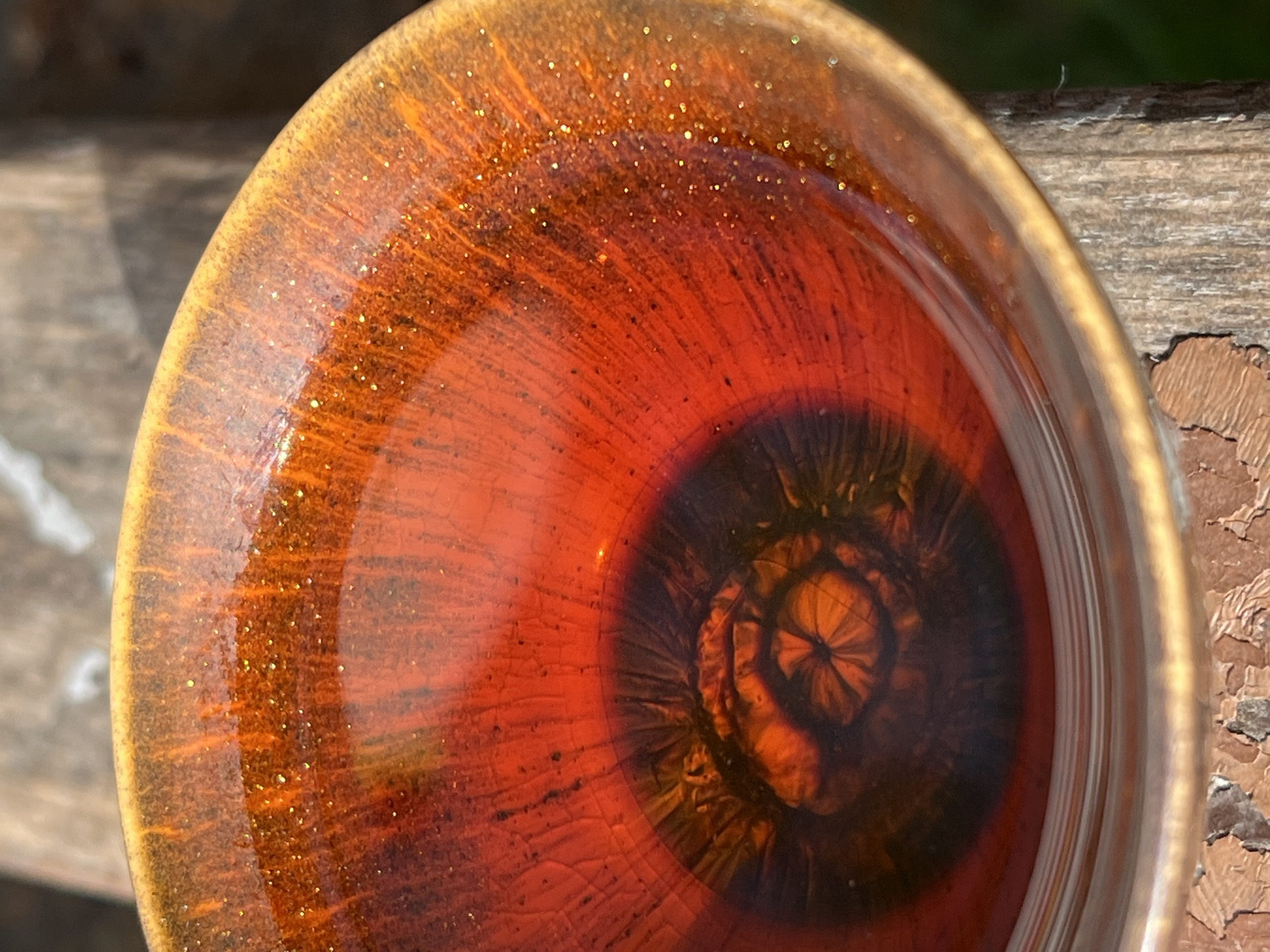 A close-up shot features a circular ceramic bowl with a glossy, textured surface. The bowl's interior displays a concentric pattern with a dark, reddish-brown core resembling a flower or abstract design, surrounded by lighter shades of reddish-brown and orange towards the outer rim. Tiny gold-colored particles are scattered across the surface, creating a glittering effect. The bowl appears to be resting on a weathered, light-brown wooden surface with peeling paint. The location in the city of Chaikovsky, Russia is not evident from the image.
