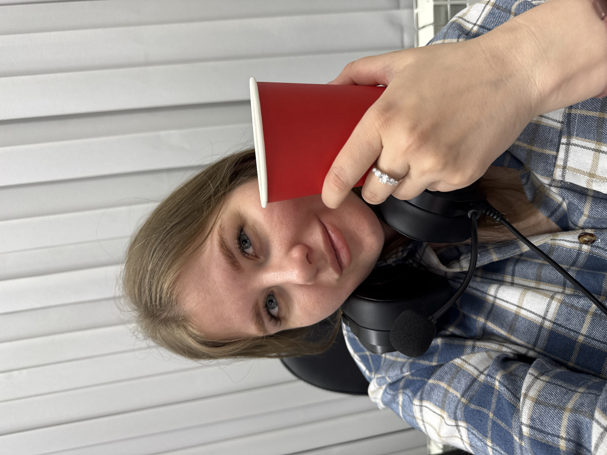 A blonde woman is holding a red paper cup with a white rim next to her face. She is wearing headphones with a microphone and a blue and white plaid shirt with brown buttons. A ring is visible on her left ring finger. The background is a white horizontal slatted blind and a wire mesh shelf. The image was likely taken in a room in Perm, Russia, given the metadata in the prompt.