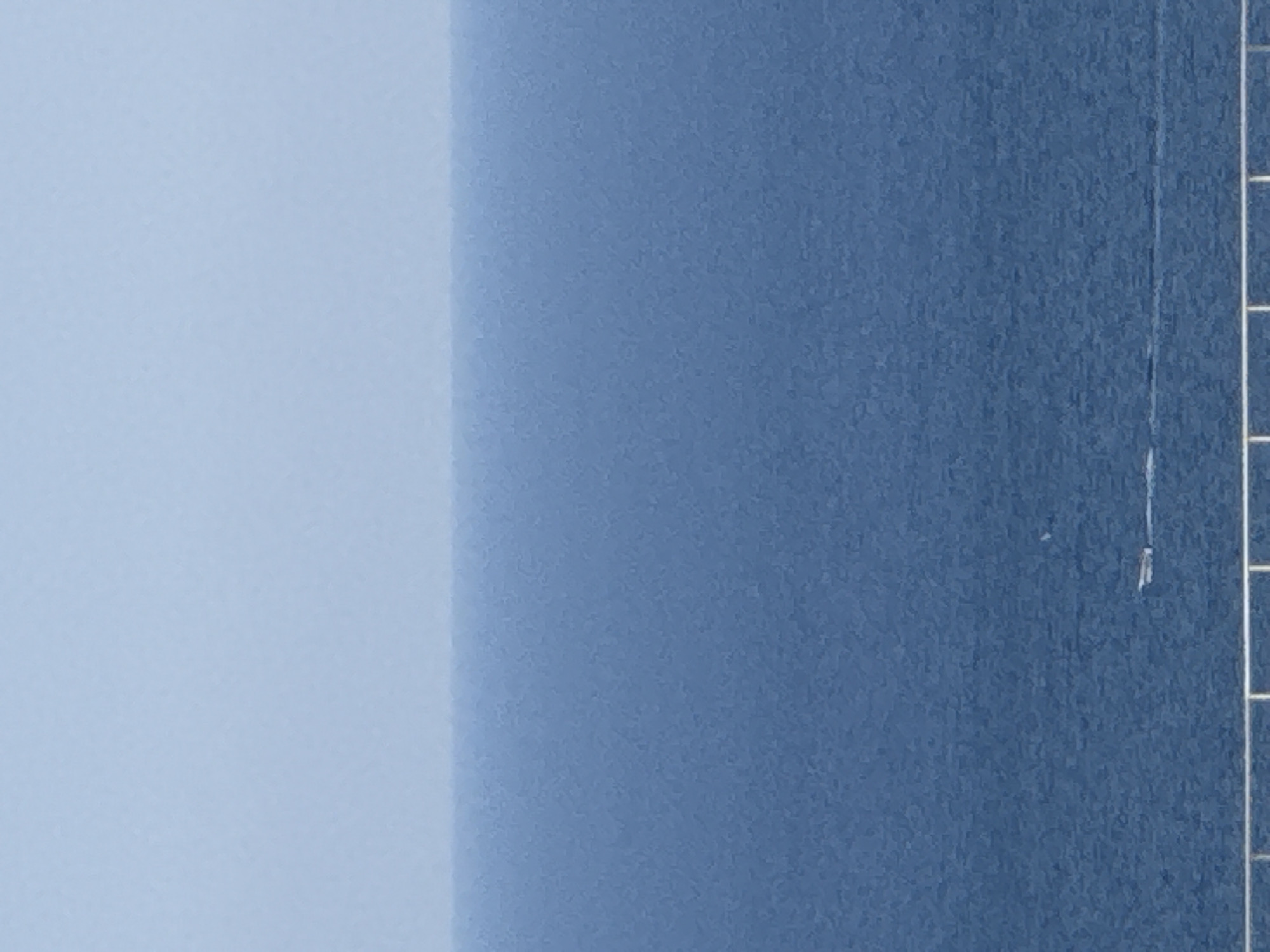 An aerial long shot captures a vast expanse of deep blue ocean under a pale blue sky. The horizon line is indistinct, with the sky and sea blending subtly. In the middle ground, a small, white boat is visible, leaving a faint white wake as it moves across the water. In the foreground, a white railing is partially visible, suggesting a viewing platform or structure overlooking the sea. The scene is serene and vast, with the distant boat emphasizing the immense scale of the ocean. The location is the coast of Boreti, Montenegro, a town known for its scenic beauty and coastal landscapes.