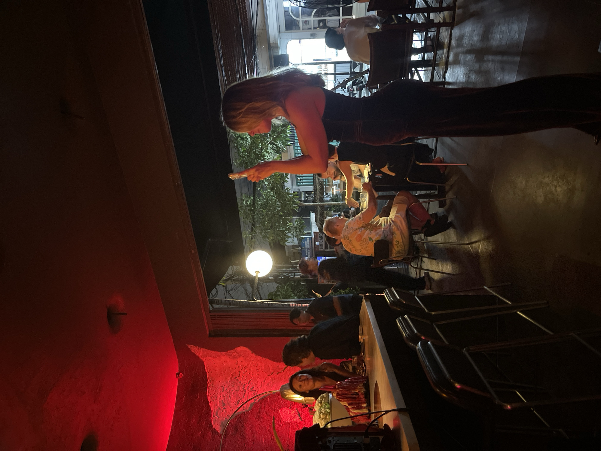 The image depicts an indoor scene at a bar, potentially in the city of Mengwi, Indonesia, illuminated by red ambient lighting and a visible round light fixture outside. In the foreground, to the right, a woman with blonde hair is standing, holding a smartphone up to take a picture or video. She is wearing a dark, strapless jumpsuit. To her left, several bar stools are visible. Further into the bar, a man with a light-colored, patterned shirt is seated at a table. Behind him, other patrons are present, including a man with dark hair and another individual with a dark shirt. On the left side of the image, a bar counter is visible, with a woman and a man standing behind it. The wall behind the bar is painted a vibrant red and features a textured, organic design. The overall atmosphere appears to be a lively social gathering.
