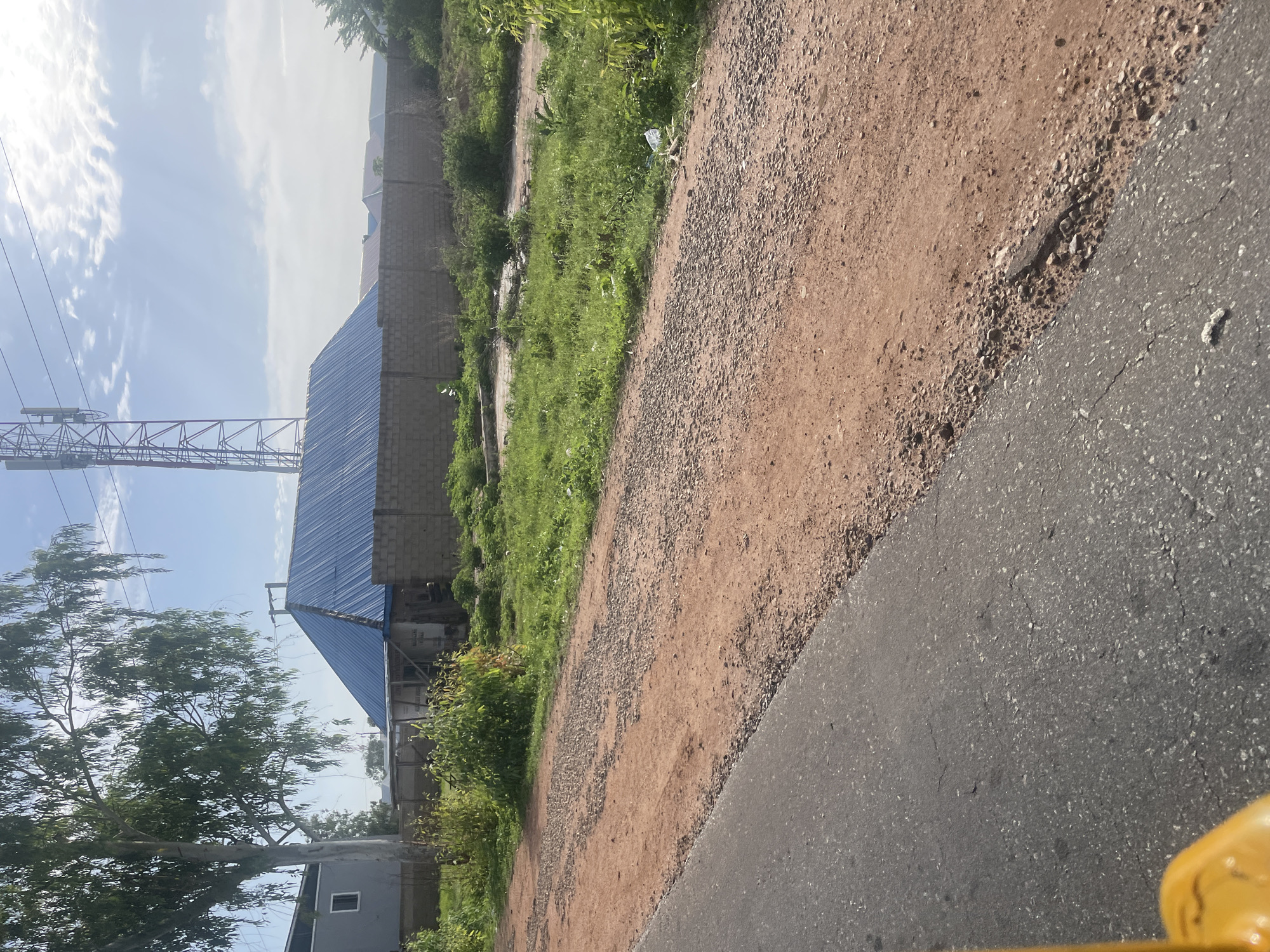 The image captures a daytime scene in Jalingo, Nigeria, focusing on a construction site bordered by a brick wall. A blue-roofed structure, partially visible, is under construction. To the left of the structure, a large tree with thin leaves stands tall, casting shadows. In the background, a telecommunications tower is prominent against a partly cloudy sky. The foreground features a section of an asphalt road with a gravel shoulder, suggesting a developing urban area. The overall environment appears to be a mix of residential and commercial development in a semi-arid climate.