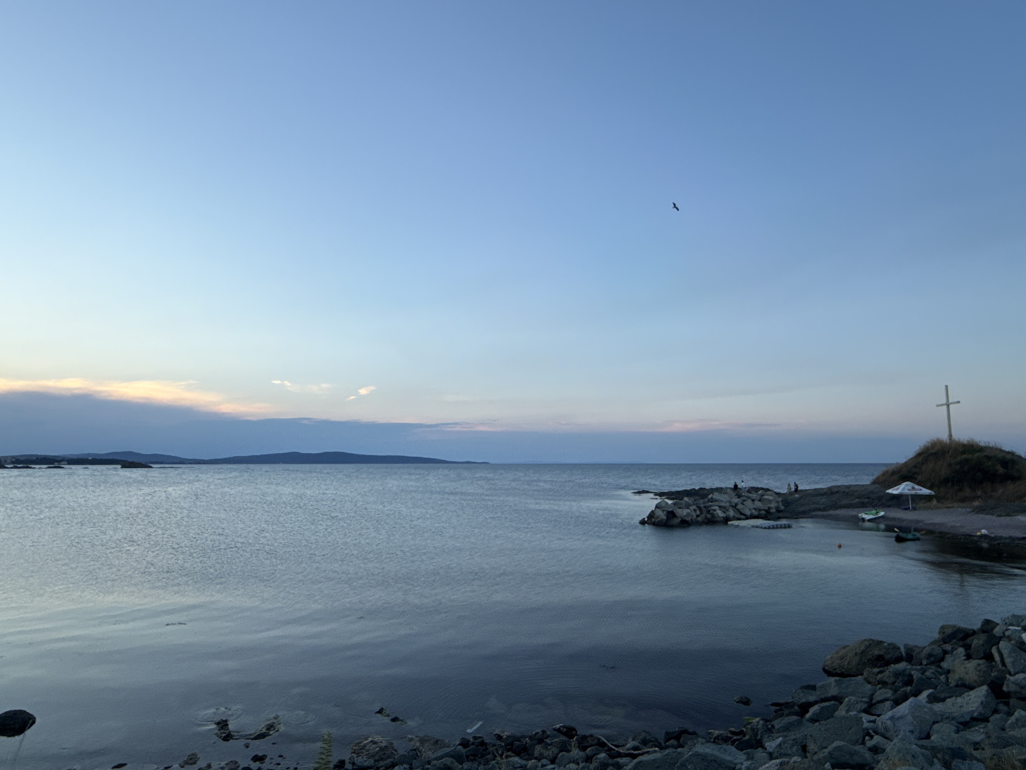 The image captures a tranquil seascape at dusk, likely in Lozenets, Bulgaria. A calm, blue body of water dominates the foreground, reflecting the soft colors of the fading sky. In the distance, a hazy, dark landmass stretches across the horizon. The right side of the image features a rocky promontory crowned with a large wooden cross. A white umbrella and two small boats are visible near the shore on the promontory. Several indistinct figures are gathered on the rocky outcroppings and near the boats. The sky above is a clear, pale blue, with a single bird silhouetted in flight. The shoreline in the foreground is lined with dark, rough stones. The overall atmosphere is one of peaceful solitude as day transitions into night.