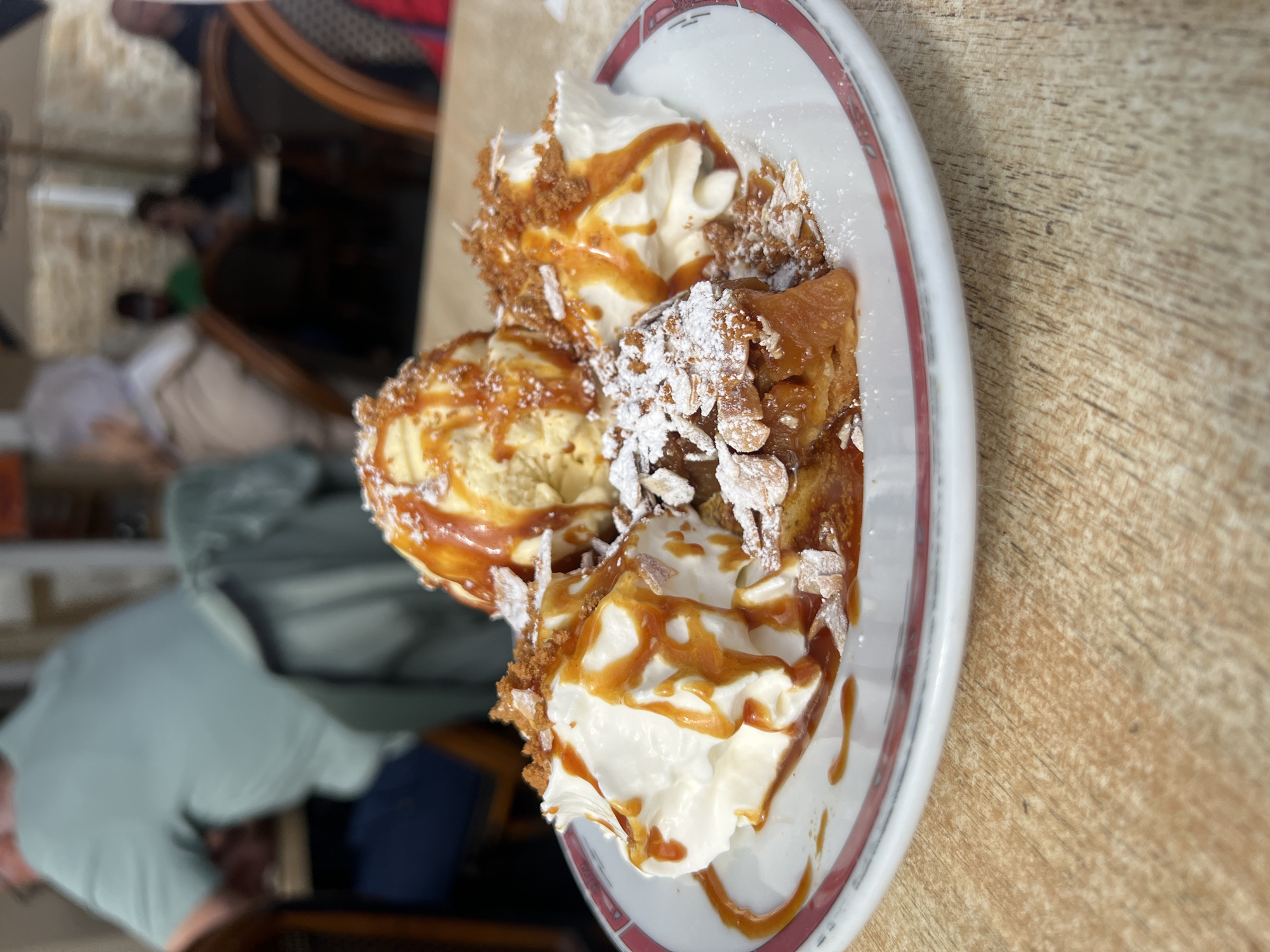 In the foreground of this close-up shot, a dessert is presented on a white plate with a red rim, resting on a wooden table. The dessert consists of a slice of apple tart, topped with whipped cream, a scoop of vanilla ice cream, and drizzled with caramel sauce. Crumbled cookies and slivered almonds are sprinkled over the whipped cream and tart, with a dusting of powdered sugar. In the blurred background, several people are seated at tables, suggesting an outdoor seating area of a restaurant. The architectural style of the buildings in the background appears to be stone, consistent with the historic nature of Le Mont-Saint-Michel, France. The overall scene captures a leisurely moment in a dining establishment within this iconic French location.