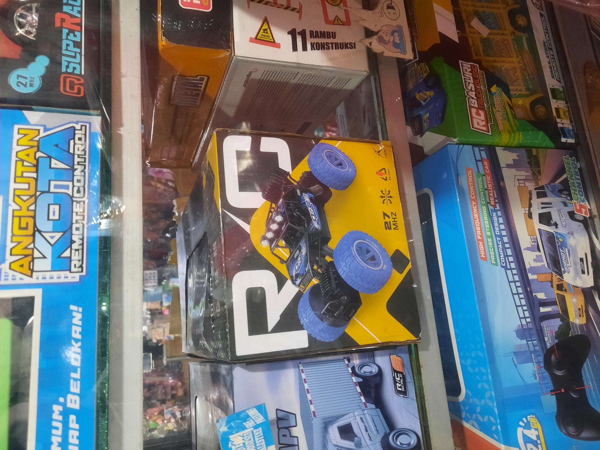 The image shows a retail display of various remote-controlled toy vehicles in their original packaging. The products are stacked on shelves in a store. In the foreground, a large box with the branding "RC BASURI TRUCK OLENG" is visible, featuring a yellow and green design with graphics of trucks and text indicating "THERES MUSIC AND LIGHTS". To the left of this, a box with a blue and white design displays a remote control and graphics of cars on a highway, with text like "HIGH FREQUENCY CONTROL" and "COMPACT DESIGN". Further to the left, another box depicts a white truck. In the middle ground, a prominent box showcases a blue and black remote-controlled car with large blue tires, labeled with "R C" in large white letters. To the right, a box with a predominantly orange and black design features the word "METAL". Above these, another box displays text related to "ANGKUTAN KOTA" and "REMOTE CONTROL" with a red and blue color scheme. The background shows the blurred interior of a store, with more products and shelves visible. The overall scene suggests a toy store or a section of a department store selling remote-controlled toys. Based on the signage visible, such as "ANGKUTAN KOTA", the location is likely Indonesia, possibly North Jakarta, given the common use of "Angkutan Kota" (city transport) as a theme for toys and local branding.