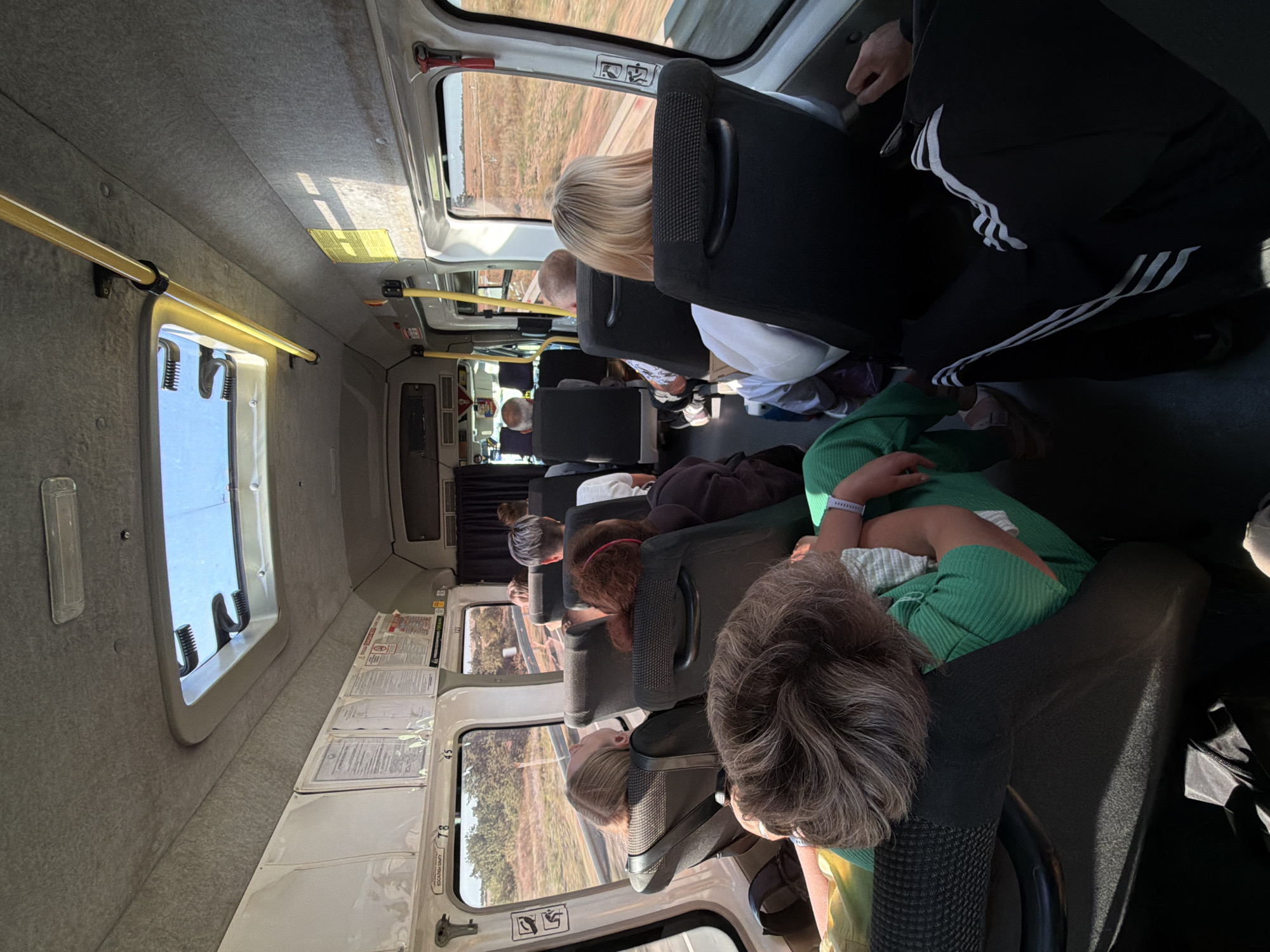The image shows the interior of a bus, likely in or around Antipovka, Russia. Several passengers are seated, facing forward. The bus has a row of windows on the right side, through which a dry, grassy landscape with some trees is visible, suggesting an outdoor or rural setting. There is a skylight in the roof, allowing natural light to enter. Yellow grab bars are installed along the ceiling. The seats are dark-colored, with some passengers leaning back or resting their heads. One passenger in the foreground is wearing a green top and appears to be sleeping or resting, with their head turned away from the camera. Another passenger in the foreground is wearing black athletic pants with white stripes. The overall atmosphere appears to be that of travel, with people journeying through the landscape.