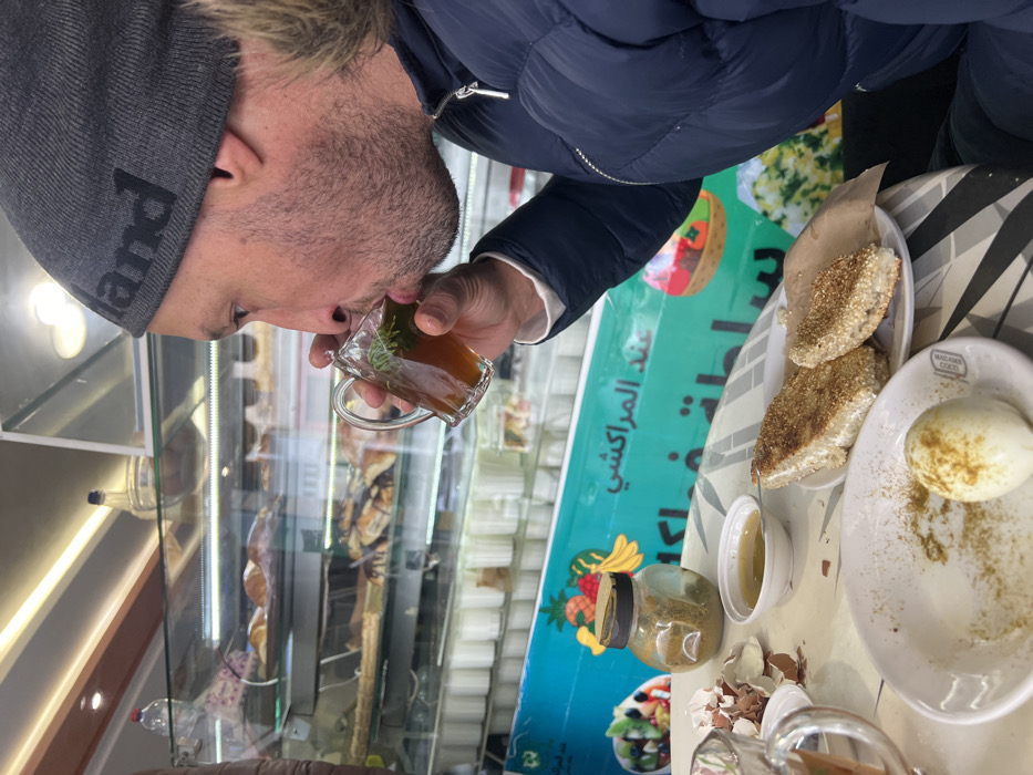 The image depicts a male individual seated at a table within a café or bakery in Meknes, Morocco. The subject, shown in profile from the right, has a short beard and is wearing a dark grey knit beanie with partially visible text and a dark blue puffer jacket with a fur-lined collar. He is actively drinking from a clear glass mug containing an amber-colored liquid with mint leaves, consistent with Moroccan mint tea.
On the table, a white plate presents a partially consumed hard-boiled egg, lightly dusted with a yellowish-brown spice blend. This plate features the text "MAHJOUBA COCK." Adjacent to it, a second white plate holds two pieces of flatbread, possibly *harcha* or *mssemen*, covered in sesame seeds and resting on a piece of brown paper. Other items on the table include a small white bowl containing a yellow liquid, likely honey or olive oil, and a small glass jar with a black lid, containing a greenish-brown powdery substance, indicative of spices. Scattered eggshells are also present on the table surface.
The background reveals a multi-tiered glass display case stocked with various baked goods, including croissants and other pastries, with rows of small white containers visible underneath. A prominent light blue banner, featuring Arabic script including "عند المراكشي" (Aind Al Marrakchi), alongside illustrations of fruits and salads, forms the backdrop. The establishment is a cafe or bakery; its specific street or neighborhood location within Meknes is not discernible from the image.