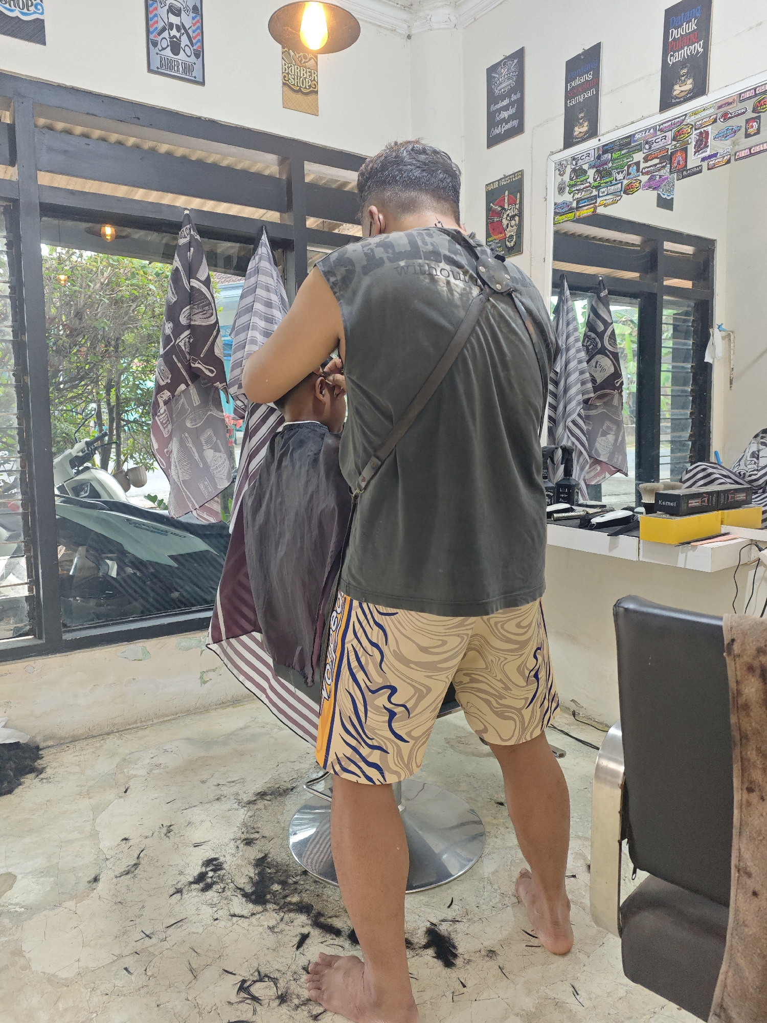 The image depicts the interior of a barber shop, likely located in Sarongan, Indonesia. A barber, wearing a dark sleeveless shirt and patterned shorts, is in the process of cutting a young person's hair. The person receiving the haircut is seated in a barber chair, draped with a cape. The floor is littered with cut hair, indicating recent activity. In the background, a mirror reflects the shop's interior, adorned with various barber shop-themed posters and stickers. A window provides a view of the exterior, where a motorcycle is visible. Various barber tools and equipment are arranged on a shelf.