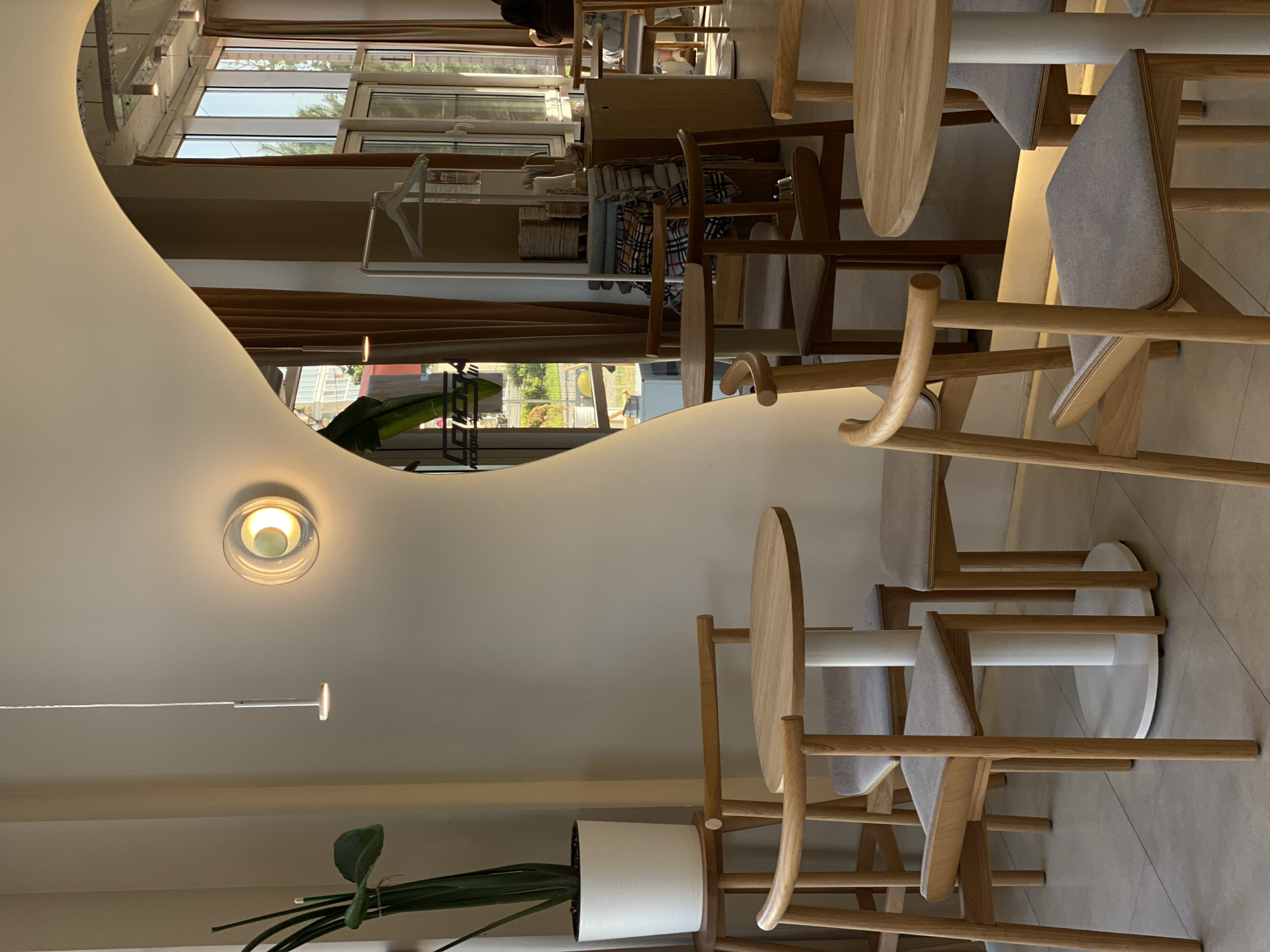 The image depicts the interior of a cafe in Yantarny, Russia, characterized by its minimalist and natural aesthetic. In the foreground, two round wooden tables with white cylindrical bases are visible, accompanied by several light-colored wooden chairs with grey upholstered seats. One table is set with a single chair, while the other appears to be awaiting patrons.
To the left, a tall, slender plant in a white pot is positioned on a wooden stool. The wall behind the plant features a decorative circular light fixture emitting a warm glow. A modern pendant light hangs from the ceiling above.
The background reveals a large, organically shaped mirror that reflects a portion of the cafe, including floor-to-ceiling windows with sheer curtains, offering a glimpse of the outdoors and the city. Through the windows, sunlight illuminates the space, highlighting a rack with folded textiles and other retail items on display. In the distance, a person is seated at a table, facing away from the camera. The overall ambiance is serene and inviting, with a focus on natural materials and soft lighting.