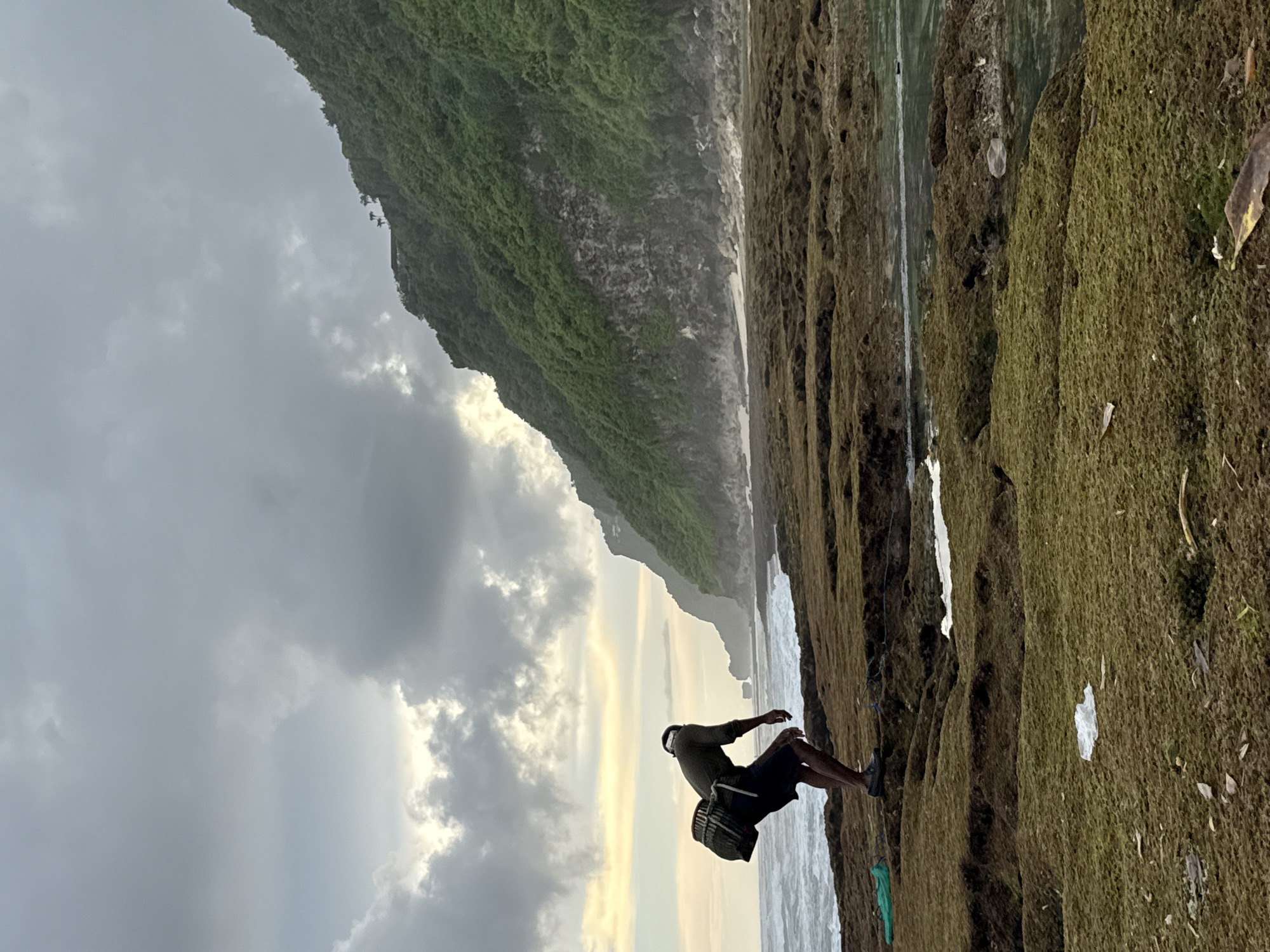 A man stands on a rocky, moss-covered shore in Pecatu, Indonesia. He is facing the ocean and appears to be bending over. The background features a cloudy sky, a green, vegetation-covered cliff, and ocean waves.