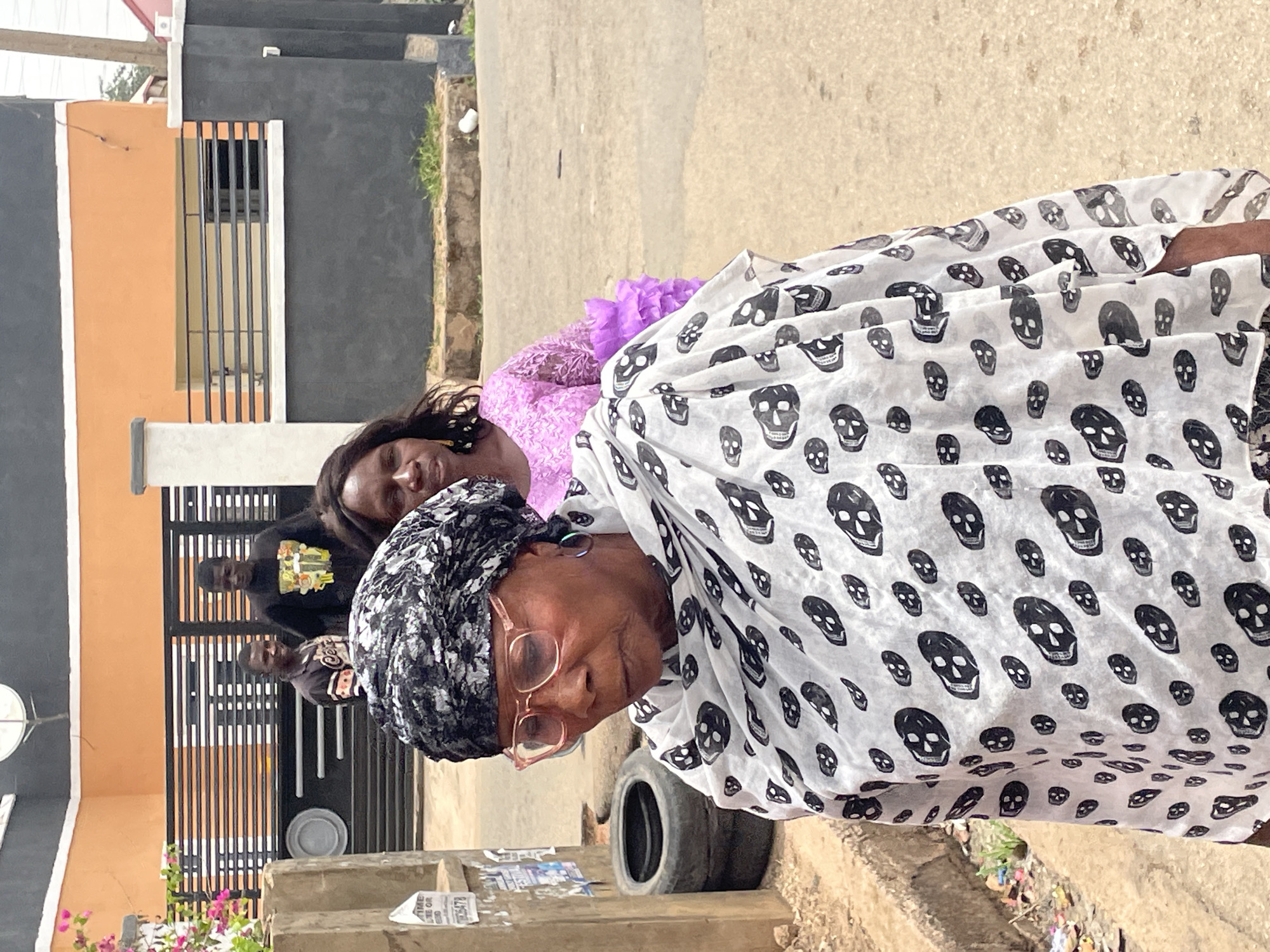 In Jos, Nigeria, the image captures a moment on an outdoor street. An older woman with glasses and a head covering is prominently featured, draped in a white shawl patterned with black skull motifs. Behind her is a younger woman wearing a purple dress, her expression serene. Further back, two men stand near a gated entrance to a building. The building is painted in a combination of orange and black, with a satellite dish visible. Street-level details include a tire and a cluttered curb with discarded items, suggesting an urban environment.