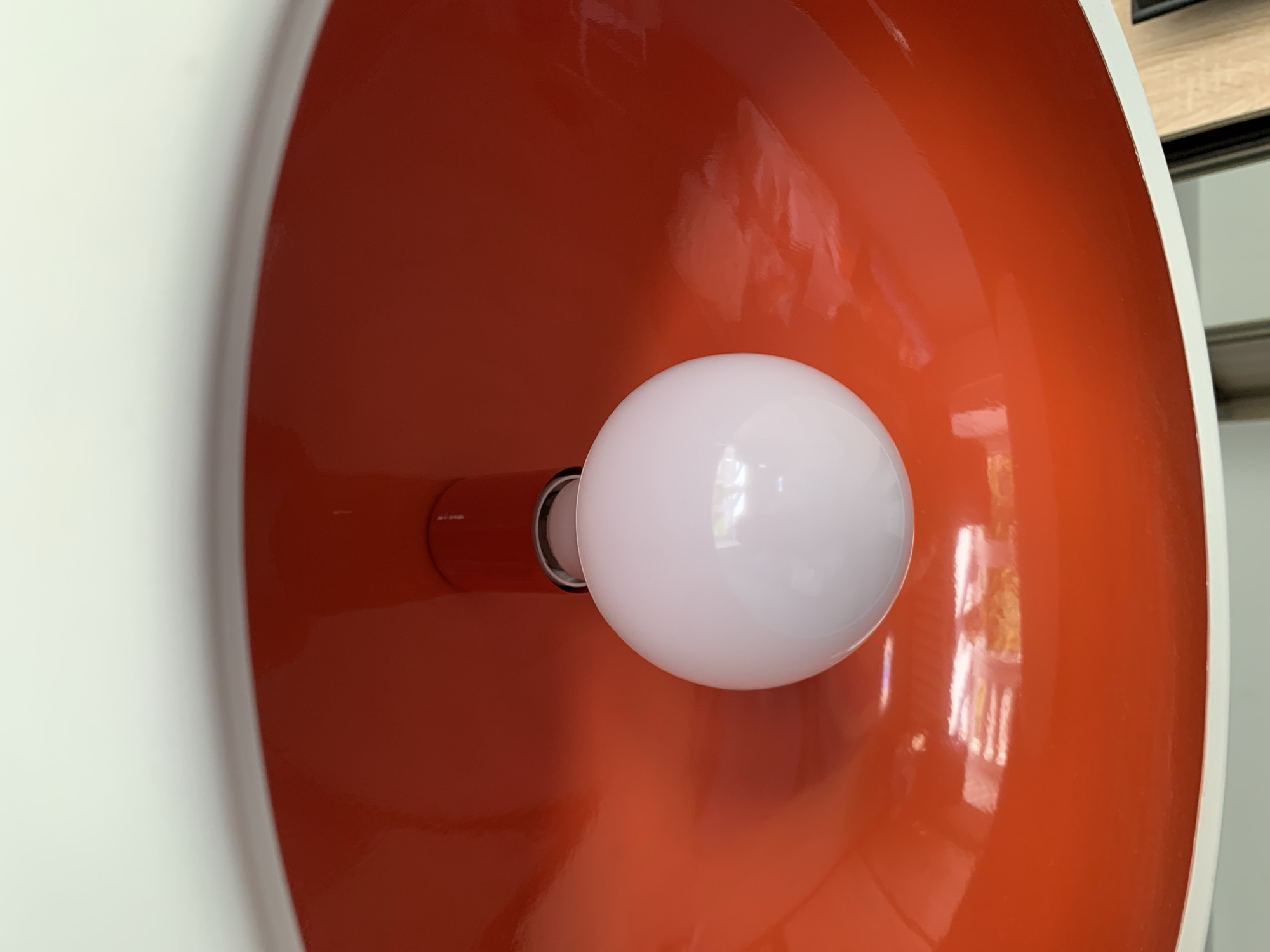 The image is a close-up, upward view of a modern, white exterior ceiling light fixture with a bright orange interior. A white, spherical lightbulb is suspended in the center, partially obscured by the orange dome. The orange interior has a glossy finish, reflecting light and showing subtle variations in color and brightness. The white exterior rim of the fixture is visible at the top of the frame. The scene appears to be indoors, possibly a hallway or room, and there is no indication of the specific location within Kapyĺ, Belarus.