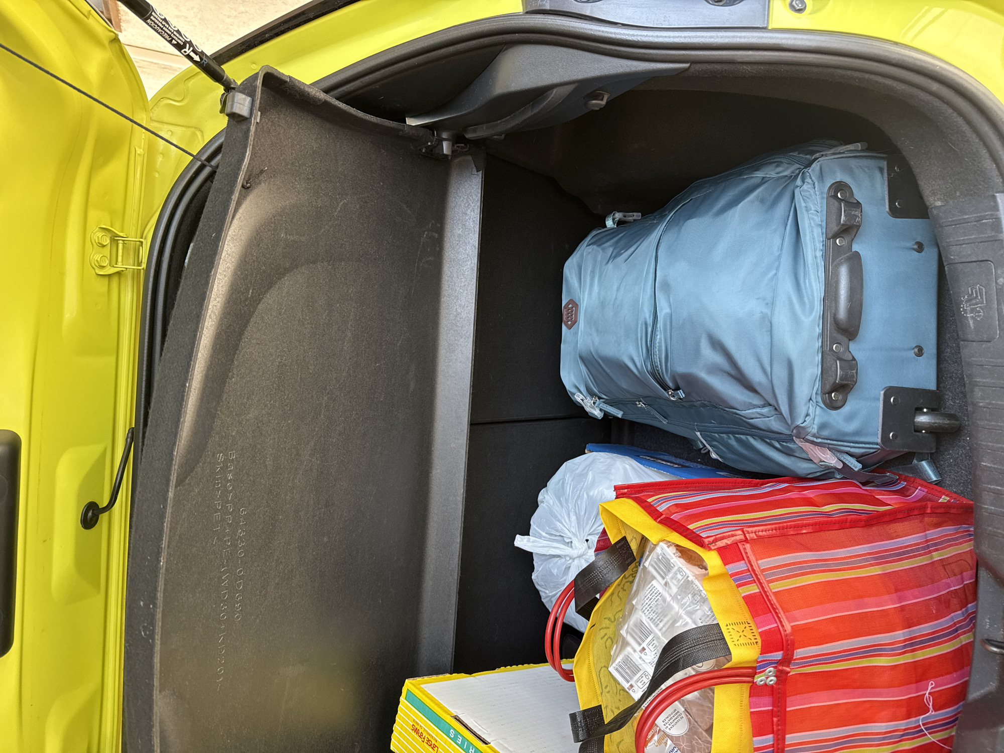 The image captures the open trunk of a bright yellow car, likely a hatchback or compact SUV, in what appears to be daytime. Inside the dark grey and black trunk, various items are packed. A prominent blue soft-sided rolling suitcase occupies the rear right section. Below and in front of it, a vibrant red, orange, and yellow striped reusable shopping bag is filled with contents, including what seem to be groceries in clear plastic packaging. Behind this bag, a white plastic bag, tied at the top, is visible. In the left foreground, a stack of yellow boxes, possibly labeled "LRSE FARMS" with a visible part reading "RIES" (suggesting "FRIES"), rests on the floor.
A gas strut, marked with "▲ PRECAUCIÓN" (CAUTION) and "NO CONTIENE" (DOES NOT CONTAIN), supports the open trunk lid. A black cord hangs from the left side of the lid. On a black plastic panel within the trunk, manufacturing information such as "64330-0D690", "Base>PP+PE-JWD30+1D201~", and "Skin>PETS" is visible. The scene depicts the process of packing or unpacking a vehicle, possibly for a trip or after a shopping outing, in Xochitepec, Mexico.