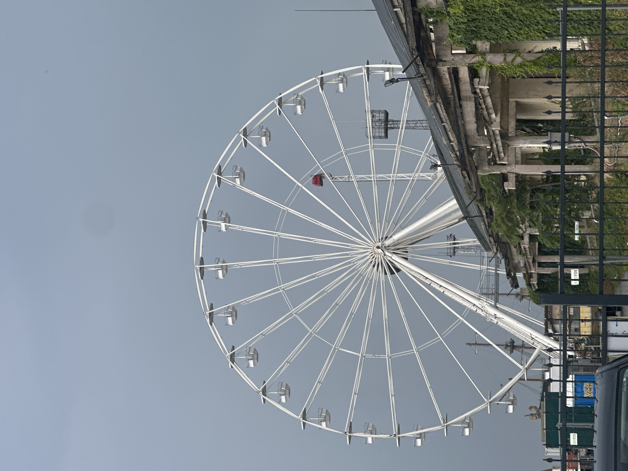A tall, white Ferris wheel dominates the center of the frame against a muted gray sky. The wheel has numerous white gondolas suspended from its spokes. In the foreground, a dilapidated building with crumbling concrete pillars and a weathered roof is partially visible, covered in lush green ivy. A black metal fence runs along the bottom of the image, obscuring some of the scene beyond. Behind the Ferris wheel, other structures, including what appears to be a ship's mast and possibly industrial equipment, are discernible. The overall atmosphere is overcast and slightly weathered.