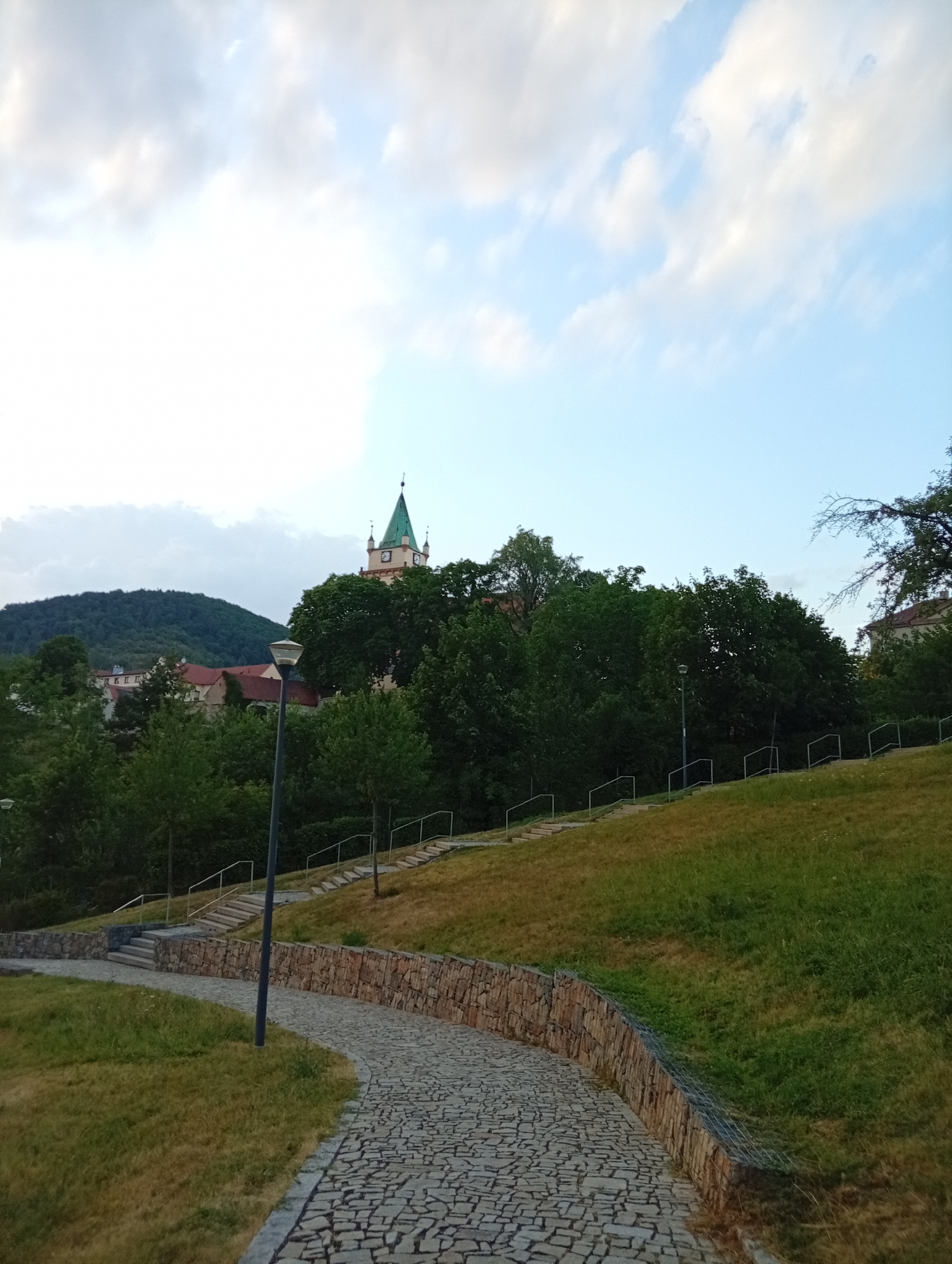 The image shows a paved pathway winding through a grassy slope with a stone retaining wall. In the background, a green-spired clock tower and red-roofed buildings are nestled among lush trees, with a wooded hill rising behind them. Several stairs with handrails ascend the slope. A street lamp stands along the pathway. The sky is partly cloudy. The scene appears to be in a park or public space within the city of Tišnov, Czechia.