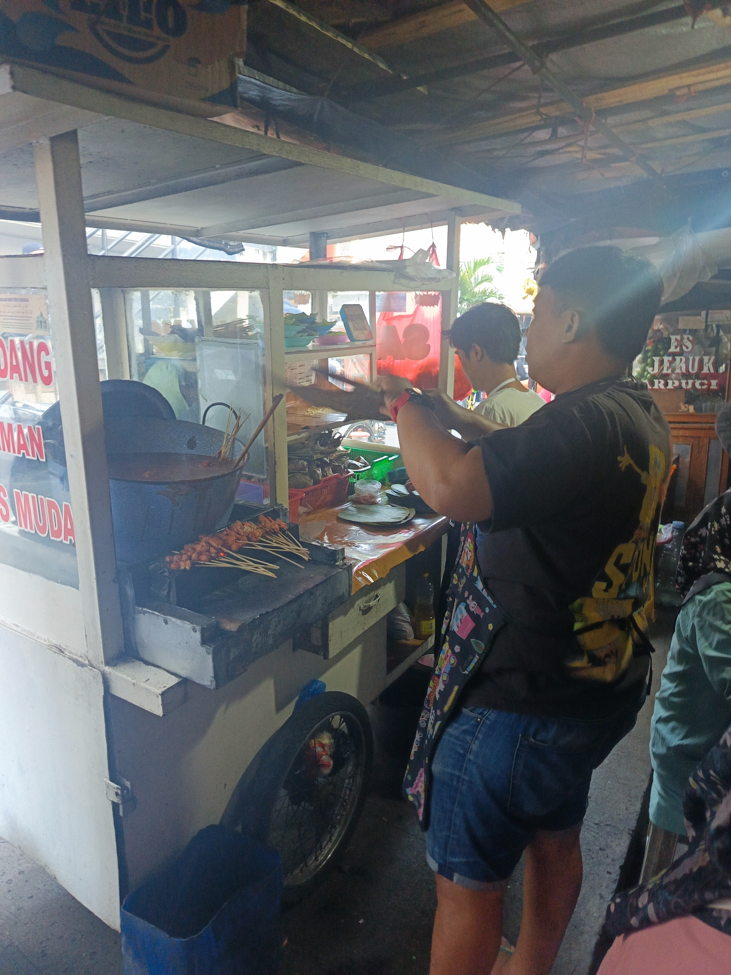 This image captures an outdoor food stall, likely in Bandung City, Indonesia, specializing in grilled skewers, possibly satay. A person, wearing a dark t-shirt and denim shorts, is seen working behind the counter, holding a spatula over a grill. Several skewers of meat are already cooking on the grill, and a large pot of sauce or broth is present. Behind the worker, another individual is partially visible. The stall is constructed with wood and glass, displaying various food items and ingredients within the glass enclosure. Signs with Indonesian text are visible on the stall. The overall environment suggests a bustling street food setting, with a focus on the preparation and sale of grilled meat skewers.
