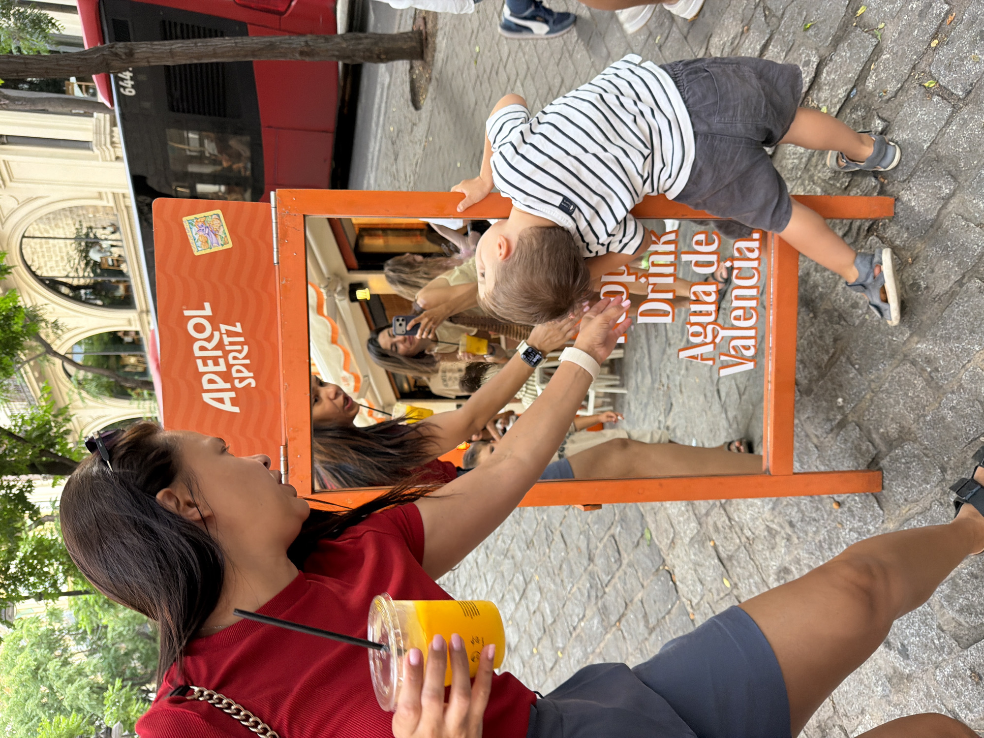 In the city of Unknown, Unknown, a woman in a red shirt and gray shorts holds a plastic cup with an orange drink and a straw. She is reaching towards a young boy who is leaning over a reflective orange stand. The boy wears a striped shirt and gray shorts. The stand has text that reads "APEROL SPRITZ", "DRINK", and "AGUA de VALENCIA". In the reflection of the stand, other people are visible, including a woman taking a photo with her phone. Behind the stand, a red bus with the number "644" is visible. The ground is paved with cobblestones, and trees and buildings are in the background.