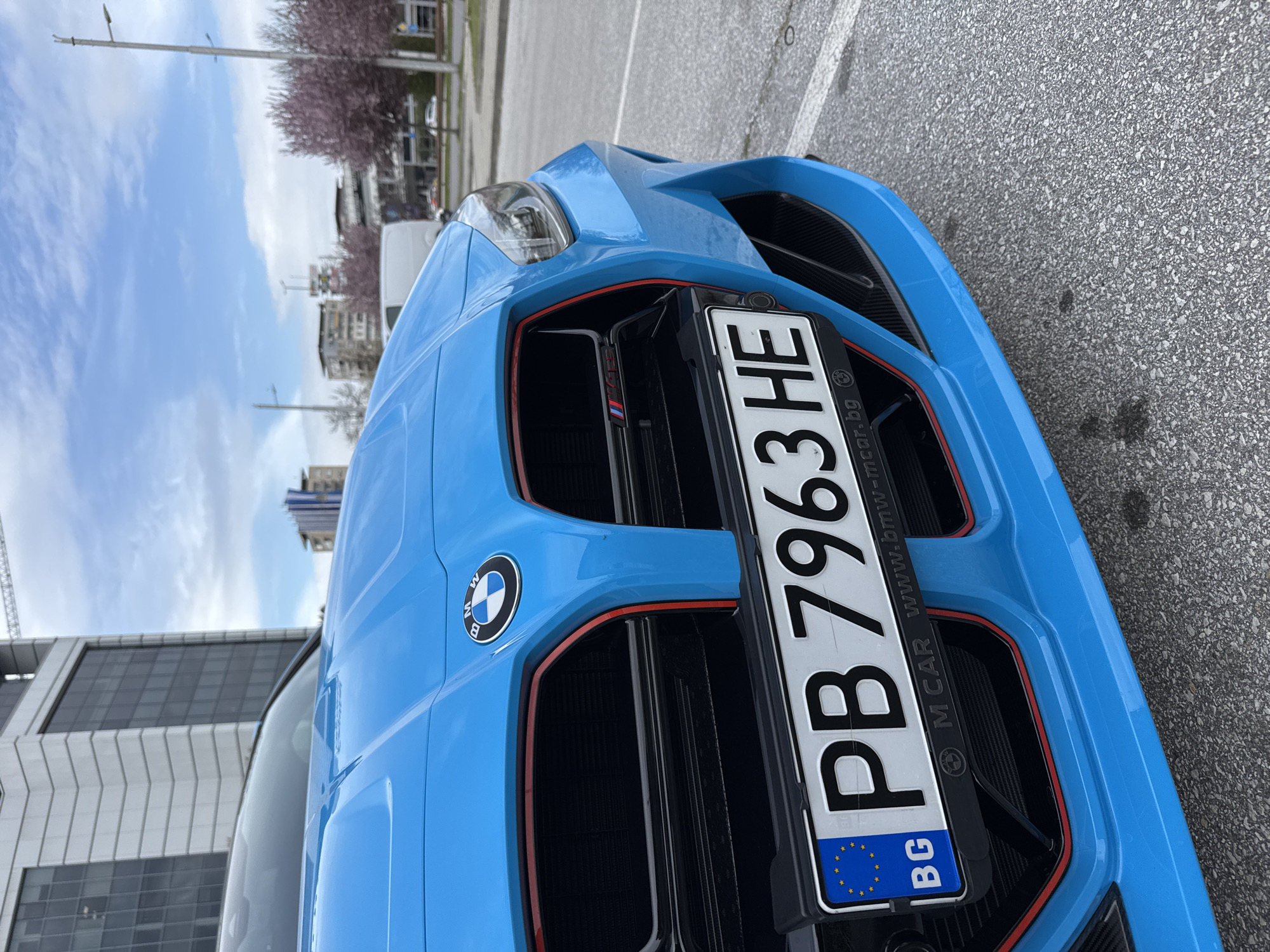 A close-up of the front of a blue BMW M3 or M4 car, positioned on asphalt in Plovdiv, Bulgaria. The focus is on the front grille, prominently displaying the Bulgarian license plate "PB 7963 HE". The M logo can be seen on the grille. The BMW badge is on the hood. The background shows urban elements, including buildings and trees, indicating an urban environment. The image is shot during the day under natural lighting.