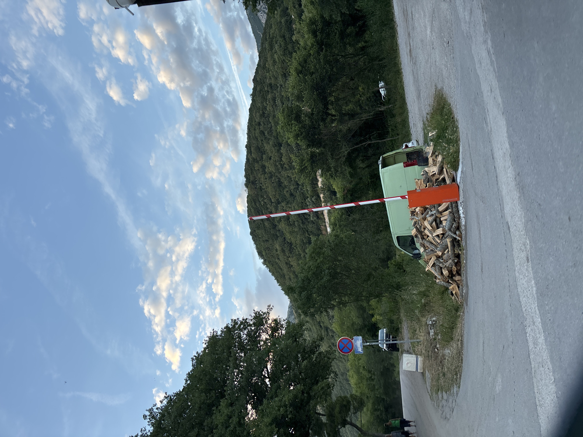 In Petrovac centar, Montenegro, a late afternoon or early evening scene depicts a paved road and a forest-covered mountainous landscape under a blue sky with scattered white clouds. In the foreground, a red and white striped barrier pole, originating from an orange base, extends diagonally upwards to the right, blocking a path. A substantial pile of chopped firewood logs is stacked around the barrier's base. Behind the firewood and barrier, a light green cargo van is parked, facing away from the viewer with its rear doors slightly open.
To the left of the barrier, a paved road curves to the left. On this road, two individuals are walking away from the viewer; the person on the left wears a dark top and bottoms, while the person on the right wears a bright green top and dark bottoms. Further up the road on the left, a circular "no parking/no stopping" sign (red circle with diagonal line on a blue background) is visible on a pole, with another indistinct rectangular sign below it. A dark-colored SUV or truck is parked behind this sign, partially obscured by foliage. In the midground, lush green trees and brush dominate, with forested mountains rising in the background under the sky, which features a faint contrail and a small dark object (possibly an aircraft or bird) in the upper left. A white vehicle is partially visible in the distant background on the right, nestled among trees.