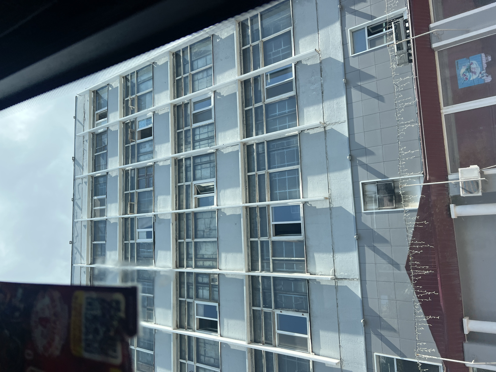 The image captures an exterior view of a multi-story building in Ulyanovsk, Russia. The building appears to be an older, functional structure with a light-colored facade, likely concrete or painted brick. It features numerous windows arranged in a grid pattern across its height, many of which seem to be closed, with some partially open or covered by blinds. The architectural style suggests a post-Soviet era construction.
The building is tall, with at least seven visible floors above the ground level. The upper floors have a lighter, more weathered appearance compared to the lower sections. The lower part of the building features a darker gray paneling and a prominent maroon-colored roof structure with decorative white string lights hanging from it, indicating a possible commercial or public entrance area. White columns support this structure. Visible on the lower levels are air conditioning units, suggesting an active or recently occupied space.
The scene is framed by dark elements on the right and the left, likely the interior of a vehicle, as indicated by the slight blur and perspective. On the far left, a portion of a sign or poster with a QR code and some red markings is partially visible. The sky above is overcast with bright, diffused light. There are no people visible in the image. The overall impression is a utilitarian urban building on a cloudy day.
