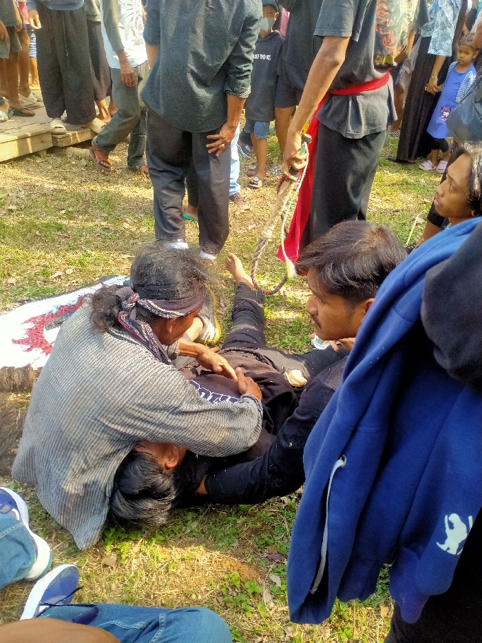 An outdoor scene in Tawangsari, Indonesia depicts a gathering around an individual lying on sunlit grass, covered in white powder/dust. A person with dark hair and a patterned bandana is kneeling, leaning over the supine individual, performing an action on their chest/abdomen area. To the right, another person with a mustache observes, wearing a black shirt with a blue hoodie draped over their arm. In the background, a person stands holding a thick rope with a loop and a red sash. Numerous other individuals, including a child in a blue dress, are visible standing around, observing the central activity from various distances, with some standing on a wooden platform.