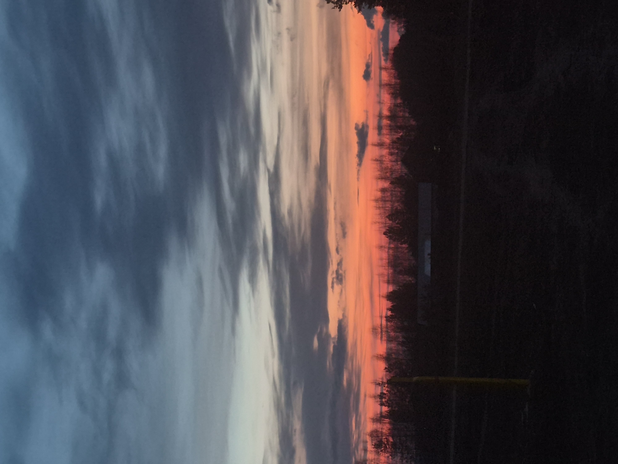 This media file presents a striking sunset over a landscape, set in what is indicated to be Chaikovsky, Russia. The image is dominated by the sky, which exhibits a dramatic gradient of colors characteristic of twilight. The upper portion of the sky is a deep, muted blue-grey, gradually transitioning into vibrant orange, red, and pink hues closer to the horizon. Wispy, elongated clouds are scattered across the sky, reflecting these brilliant colors.
Along the horizon, a dark, silhouetted line of dense trees and what appear to be distant structures or buildings is visible, creating a sharp contrast against the radiant sky. The foreground below this silhouette is plunged into deep shadow, making details difficult to discern. However, a faint, thin horizontal yellow line or object can be seen extending across the lower-right portion of this dark foreground.
The scene captures the tranquil beauty of late evening, with the sun having just set or being very low on the horizon, casting a warm glow across the clouds. No people or specific activities are visible, emphasizing the natural landscape. The weather appears clear enough for the sunset colors to be prominent, with scattered cloud cover adding texture to the sky. There is no visible text in the image.