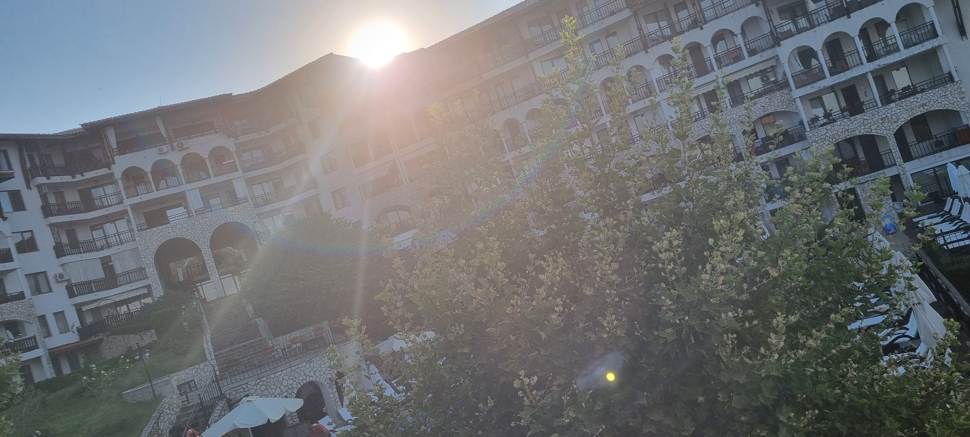 The image depicts a sun-drenched, multi-story hotel building with a curved facade, likely situated in a coastal resort area. The architecture features numerous balconies with dark railings, arched windows, and stone accents. Lush green trees partially obscure the lower levels of the building. In the foreground, a bright sun causes lens flare and washes out parts of the scene. To the right, lounge chairs and white umbrellas suggest a poolside or beach area. To the left, a white umbrella is visible, along with hints of stairs and greenery. The overall impression is of a sunny day at a resort, possibly in Sveti Vlas, Bulgaria, given the context provided, though the image itself does not explicitly confirm the location.