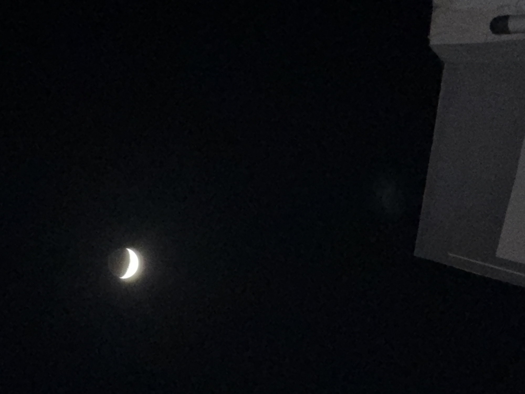 The image depicts a crescent moon in a dark sky, likely at night. In the upper right corner, there is a white, rectangular structure, possibly part of a building. The location is assumed to be Kuta Selatan, Indonesia, but there are no location-specific markers visible in the image. No people, interactions or actions are visible in the image.