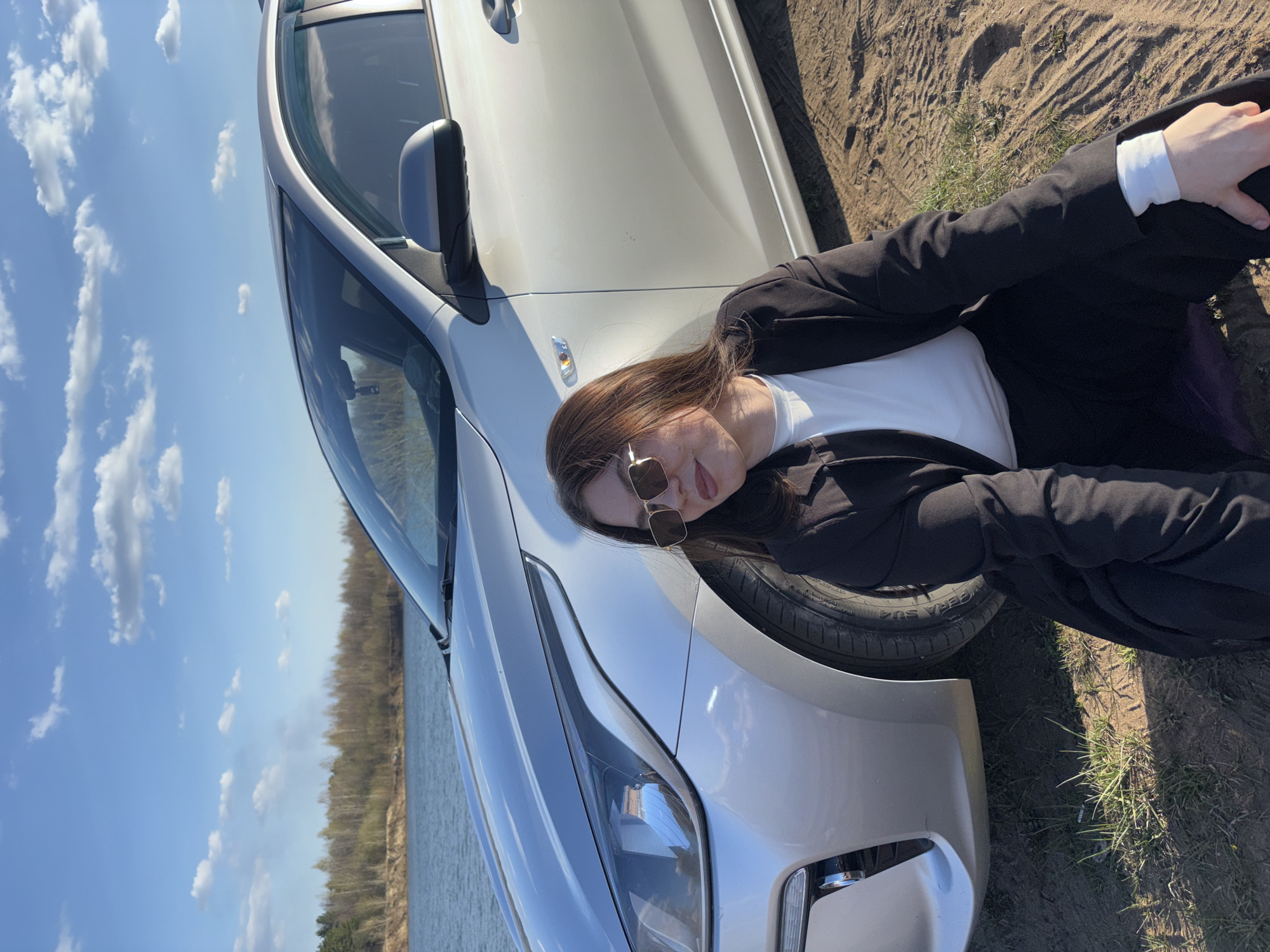 A woman in a white shirt, black blazer, and sunglasses is lying on the hood of a silver car. The car is parked on a dirt surface near a body of water. The sky is blue with scattered clouds. The location is near a body of water in Zaosinovo, Russia.
