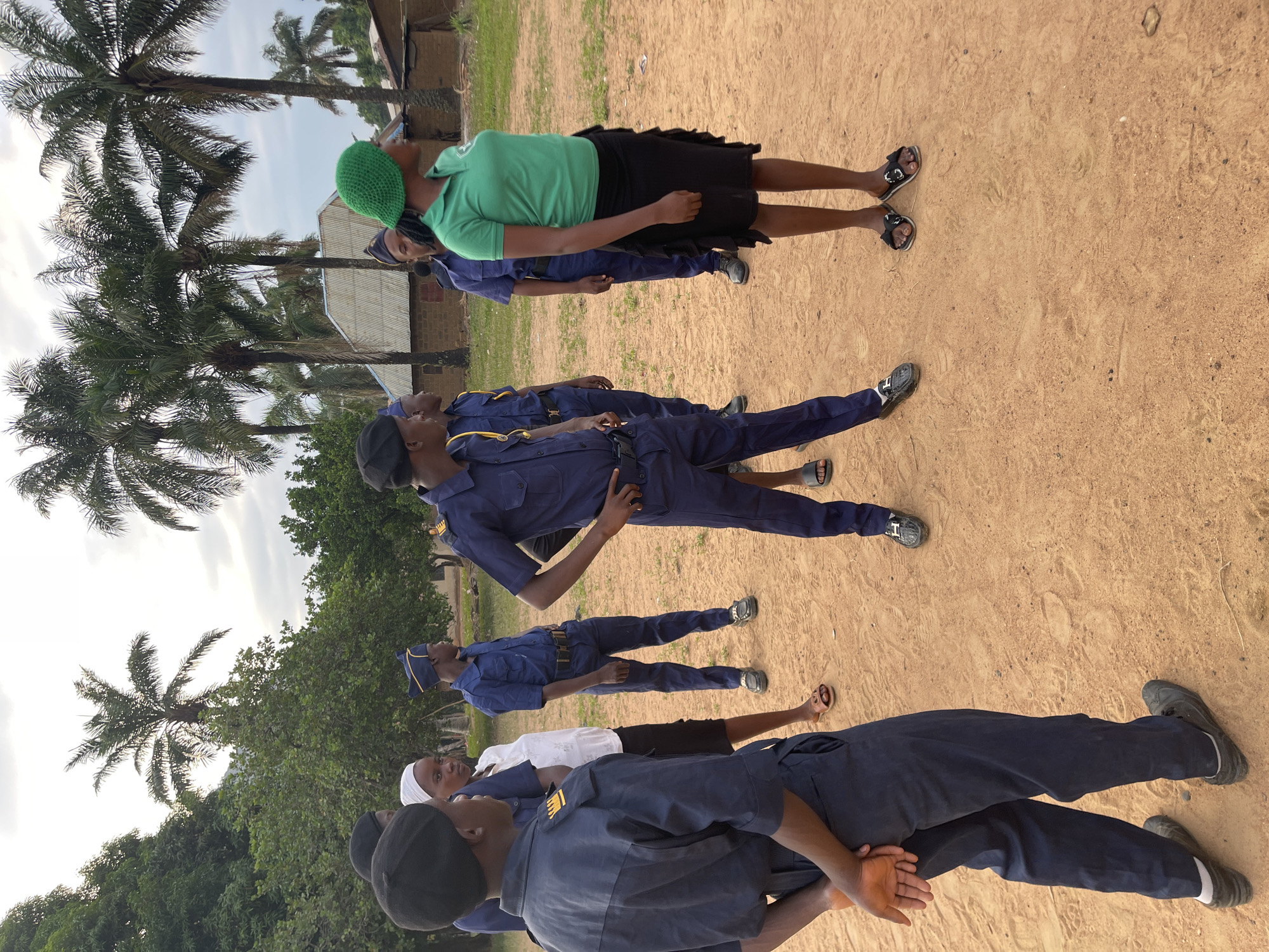 A group of individuals are standing in rows on a dirt field. Most are wearing dark blue uniforms with yellow accents, likely indicative of a security or training group. One individual stands apart, wearing a green shirt, black skirt, and green knitted hat. Palm trees are visible in the background. The location is likely a training area or field in Tabu, Nigeria.