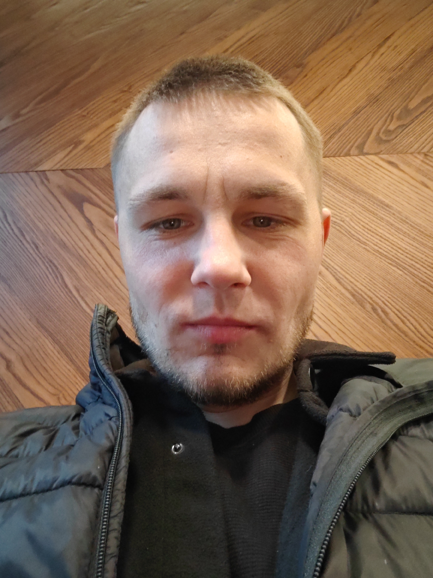 A man is seen in a close-up, straight-on shot. He has short, light brown hair, stubble on his face, and light-colored eyes. He is wearing a dark-colored jacket over a dark sweater. The background is a wood-paneled wall. The location cannot be determined as Hrodna, Belarus based on the image.