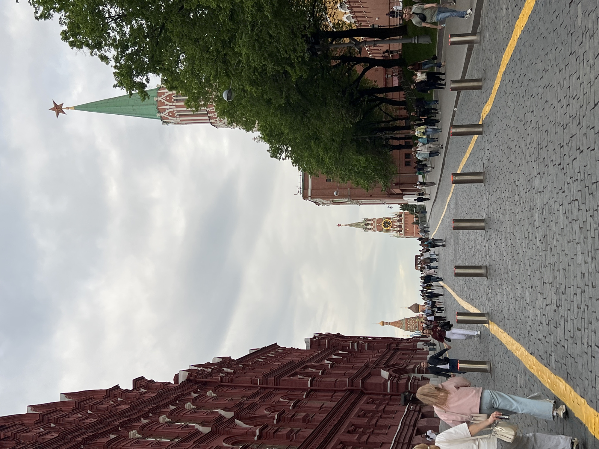 The image shows a view in Moscow, Russia, near the Kremlin. It features several towers of the Kremlin and the adjacent buildings along Red Square. Pedestrians are walking along a stone-paved walkway marked with a yellow line. There are metal bollards along the walkway. The sky is overcast. The architecture is traditional Russian style with red brick and ornate details. Trees are present between the towers.