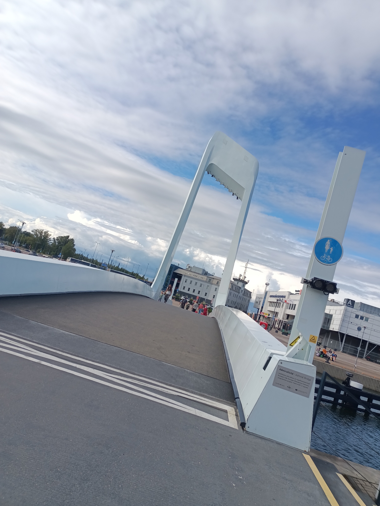 The image shows the Admiral's Bridge in Tallinn, Estonia. It is a modern, white pedestrian and cyclist bridge spanning a body of water, likely a harbor or canal, given the presence of docked boats and industrial-looking buildings in the background. The bridge has a distinctive arching structure.
On the bridge, there are several people walking, some are dressed in casual clothing. A blue and white circular sign indicating pedestrian and cyclist use is visible on a white pillar of the bridge. The road surface of the bridge is asphalt with white markings.
The background features a cityscape with multiple buildings, some appearing to be commercial or public structures, with a clear sky above dotted with white clouds. Trees and streetlights are also visible along the distant waterfront. The water appears dark and calm. A sign on the side of the bridge provides information in Estonian and English, identifying it as the "Admiral Bridge" and mentioning its connection to an EU funded project.