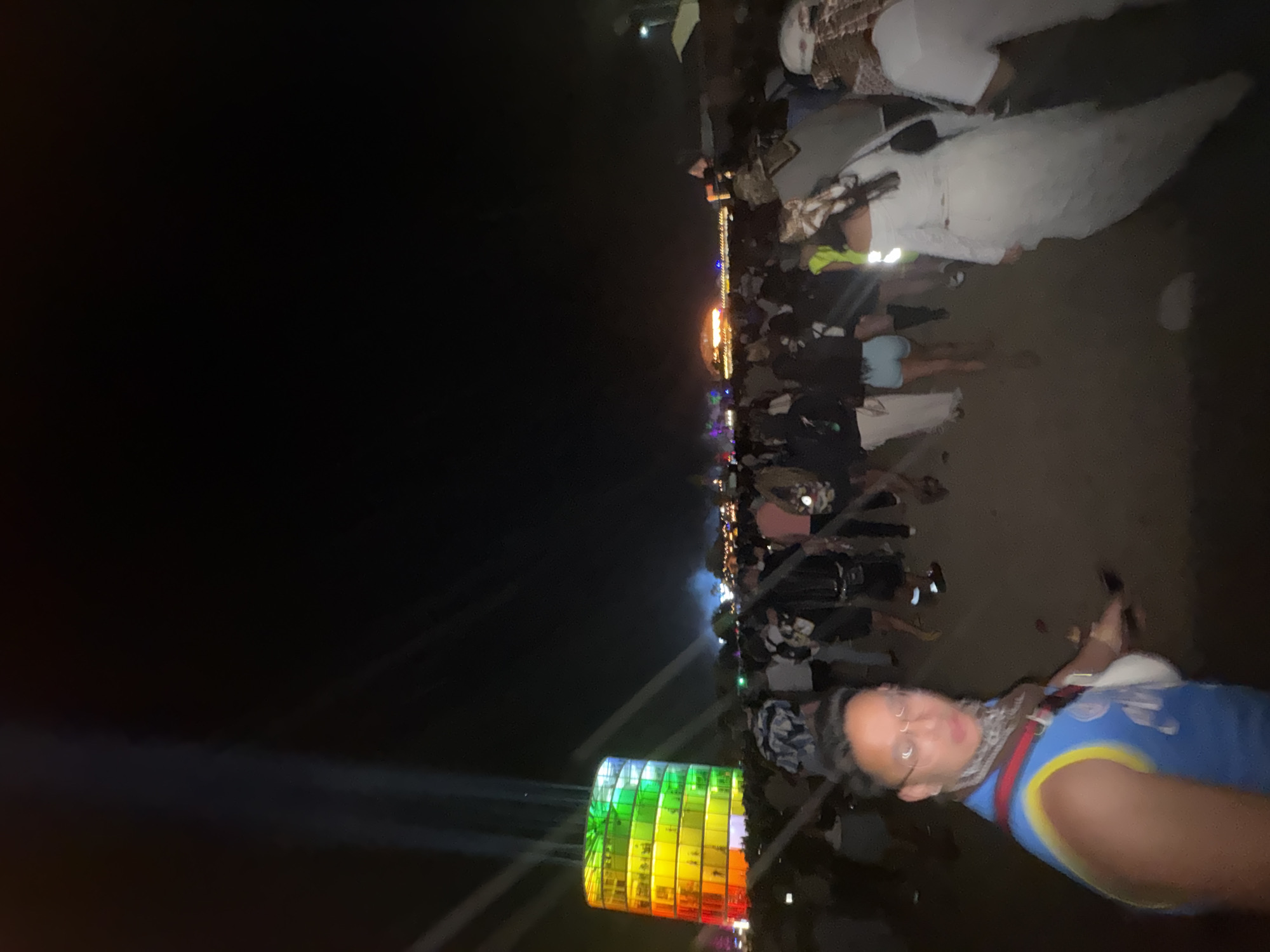 At night in Indio, United States of America, a person in a blue and yellow tank top is holding a camera and taking a selfie. Behind the person is a crowd of people, and in the distance is a large, colorful structure with lights shining from it.