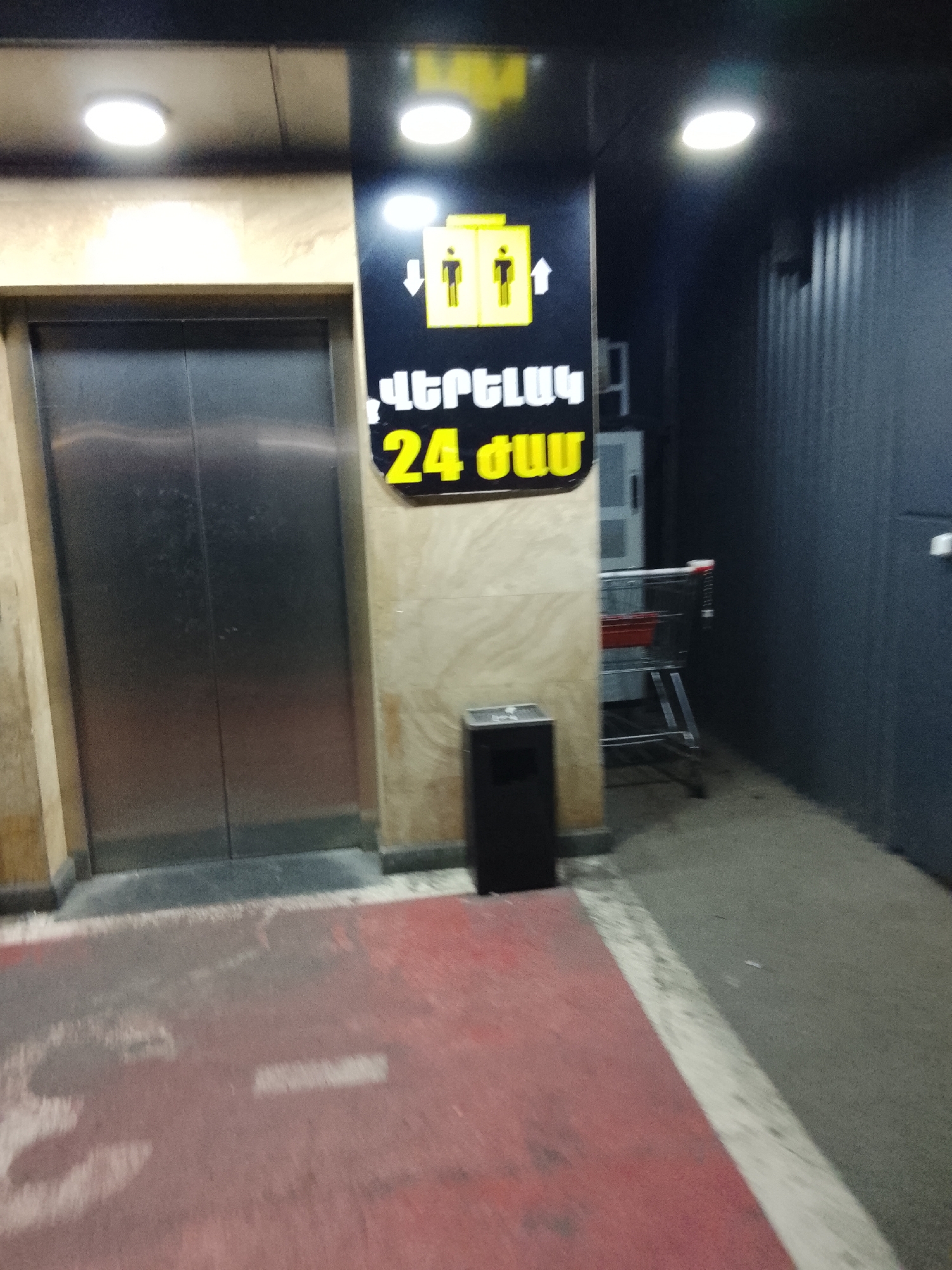 The image captures an elevator entrance area, likely within a commercial building or supermarket, illuminated by artificial ceiling lights, suggesting it's either indoors or nighttime.
On the left, a stainless steel elevator door is visible, framed by light brown, possibly tiled, walls. Above the elevator, two circular ceiling lights cast bright glows. To the right of the elevator, a prominent black sign is affixed to the wall. This sign features two yellow elevator icons with directional arrows: one pointing upwards and one pointing downwards. Below these icons, yellow Armenian text is displayed: "ՎԵՐԵՎ" (Verev), which translates to "UP," and "24 ԺԱՄ" (24 Zham), which means "24 HOURS" or "24/7."
At the base of the wall beneath the sign, a black, rectangular trash bin or ashtray is situated. Further to the right, a metallic shopping cart with a red plastic basket is parked next to a dark, vertically ribbed or corrugated metal wall. Another circular light is visible above this section.
The foreground features a red painted floor that appears worn, with some lighter markings. This red surface transitions into a grey concrete or paved area extending towards the right and under the elevator. The overall lighting is artificial and bright, highlighting the elevator and the sign.