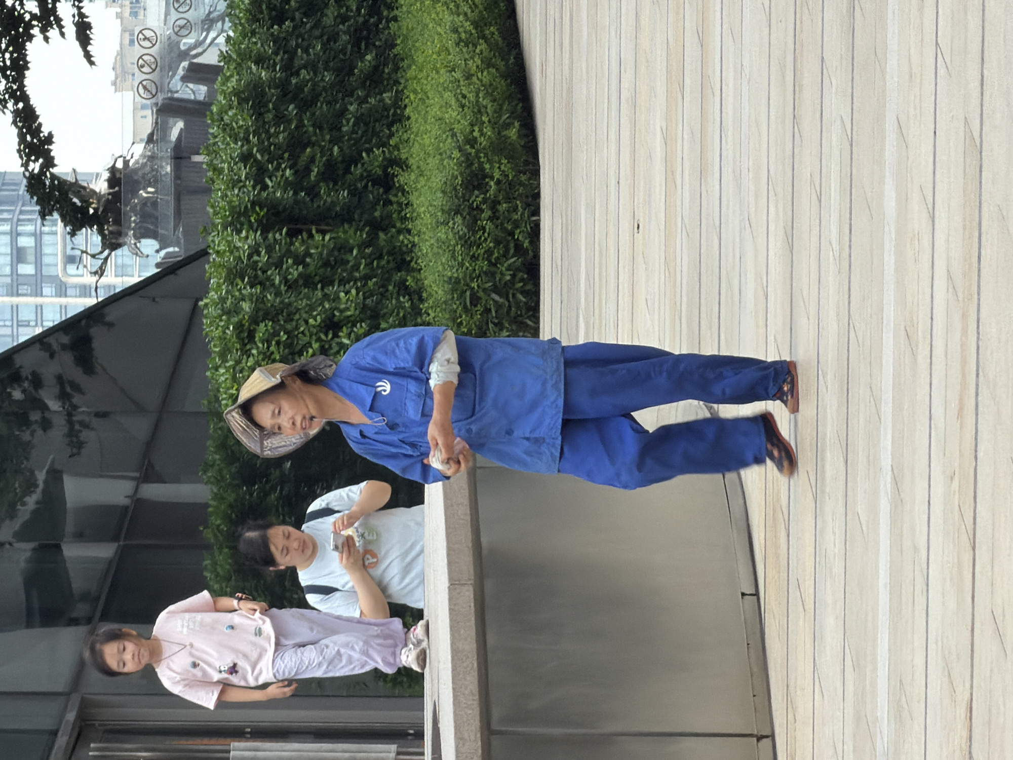 In Yuzhong District, China, a woman in a blue uniform and hat walks on a wooden boardwalk. The woman is holding a small white object in her right hand. She has a microphone clipped to her collar. To her left, a young girl in a pink t-shirt and white pants stands looking forward. Behind the girl, another woman in a light blue t-shirt holds a smartphone. The background features a building with dark, reflective windows and lush green bushes. The overall scene appears to be an outdoor public space, possibly a park or a commercial area.