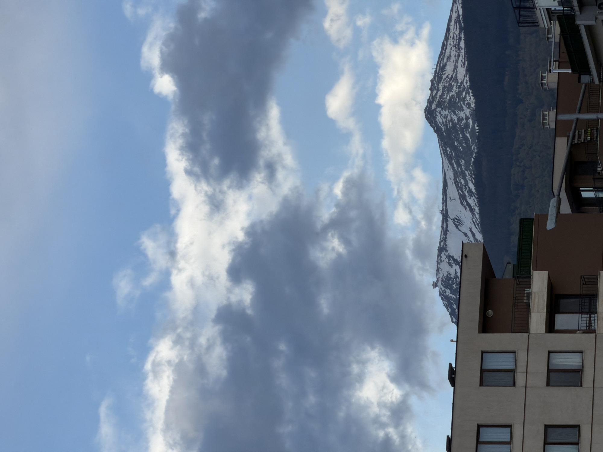 The image shows a view of a building in Sofia, Bulgaria, with a snow-capped mountain in the background. The sky is blue with some gray clouds. The building has a light-colored facade and several windows. There are also balconies on the building. The mountain is partially covered in snow. The image is rotated.