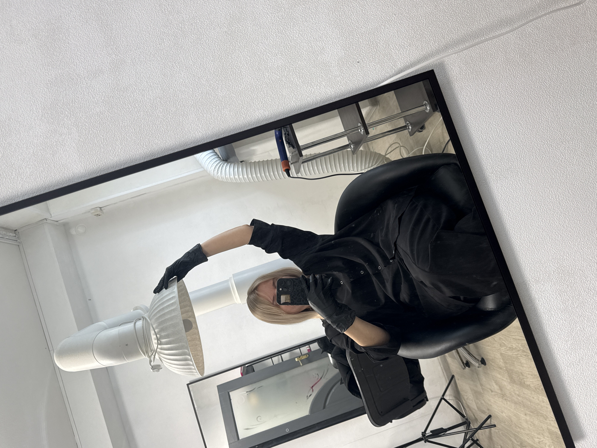 A selfie reflected in a mirror shows a person with blonde hair, wearing black gloves and a black outfit, holding a smartphone. The person is sitting in a black chair. Above and to the left of the person is a white ventilation system. The mirror's reflection reveals a window or door and a white tiled floor. The mirror is framed in black and partially reflects a white wall. The city of Chaikovsky, Russia, cannot be confirmed from the image.