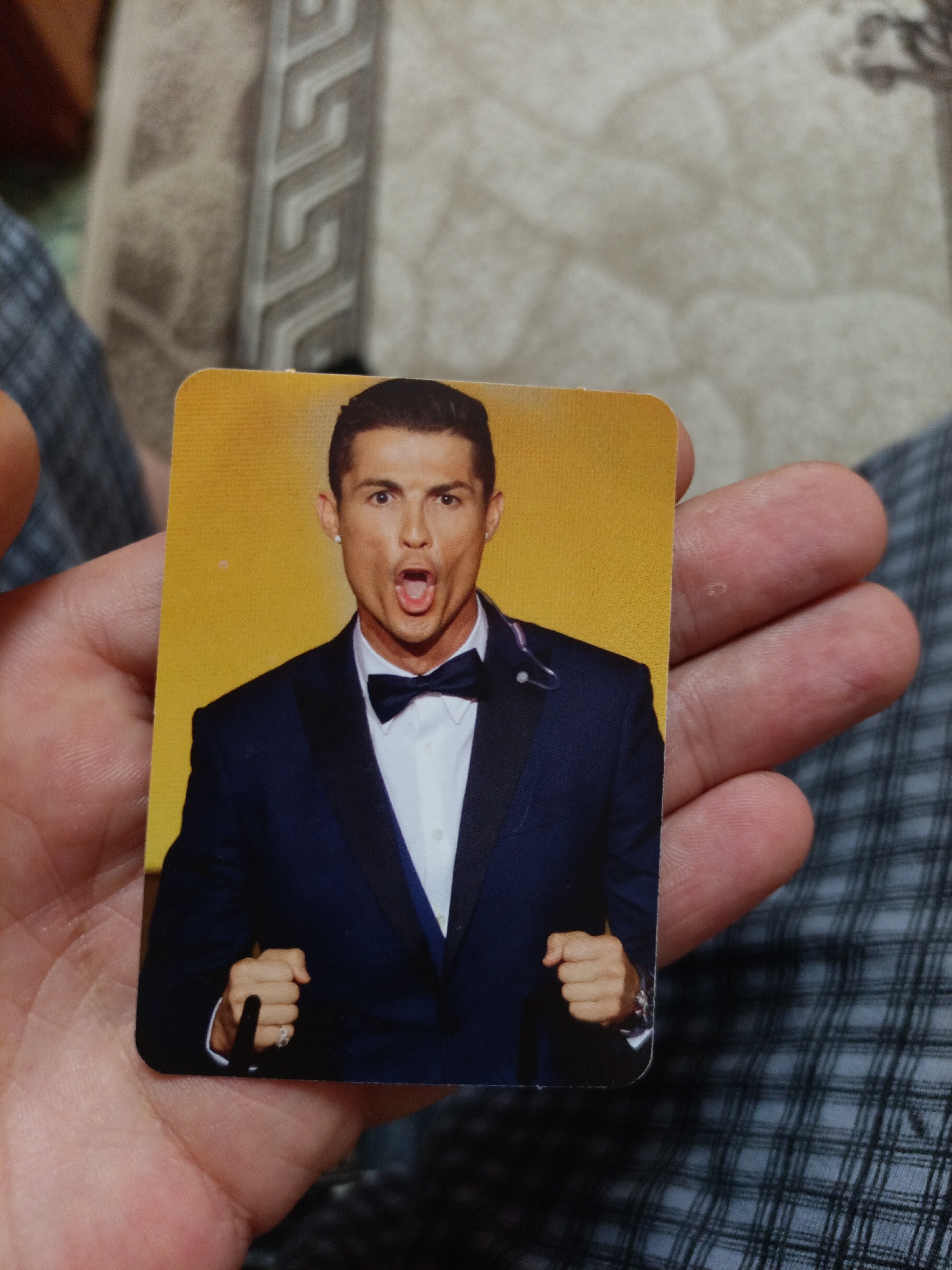 A person's hand holds a rectangular card featuring a portrait of Cristiano Ronaldo. He is wearing a dark blue suit with a white dress shirt and a black bowtie. Ronaldo's mouth is open in a yell or shout, and he is wearing a single white earring. The background of the card is a solid yellow. The hand holding the card has a ring on the ring finger. The background of the image shows a patterned fabric, possibly upholstery or clothing. The image does not contain any information about the city of Nyagan, Russia.