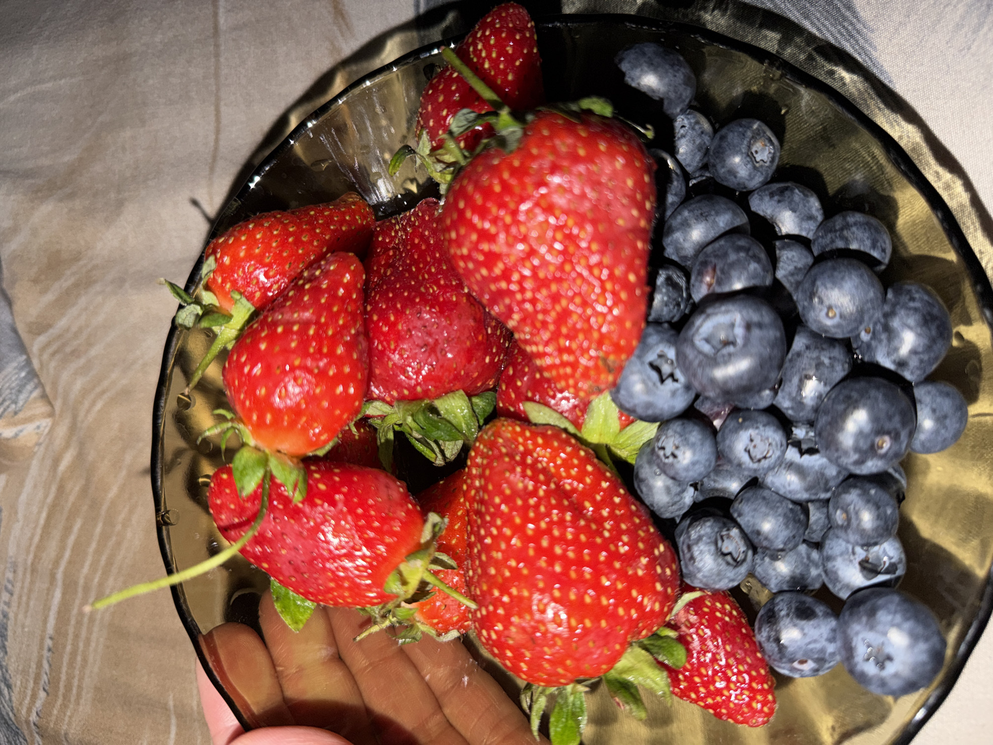 A bowl of fresh strawberries and blueberries is being held in someone's hand. The bowl is transparent and sits on a neutral-colored fabric. The strawberries are red and appear ripe with green stems attached. The blueberries are a dark blue-purple color. The person's hand holding the bowl has tan skin. The image appears to be taken indoors under artificial lighting. The location is not identifiable from this image and there's no indication it is in Moscow, Russia.