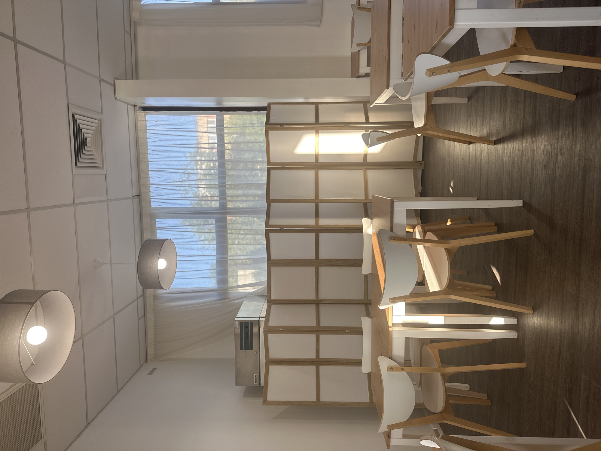 The image shows an interior space, possibly a restaurant or cafeteria, located in Ramonville-Saint-Agne, France. The room features a window covered with a sheer white curtain, next to a room divider and an air conditioning unit. There are wooden tables and white chairs arranged in rows, with light-colored wooden legs. Two pendant lamps with gray shades are suspended from the ceiling. The floor is a dark brown wood.