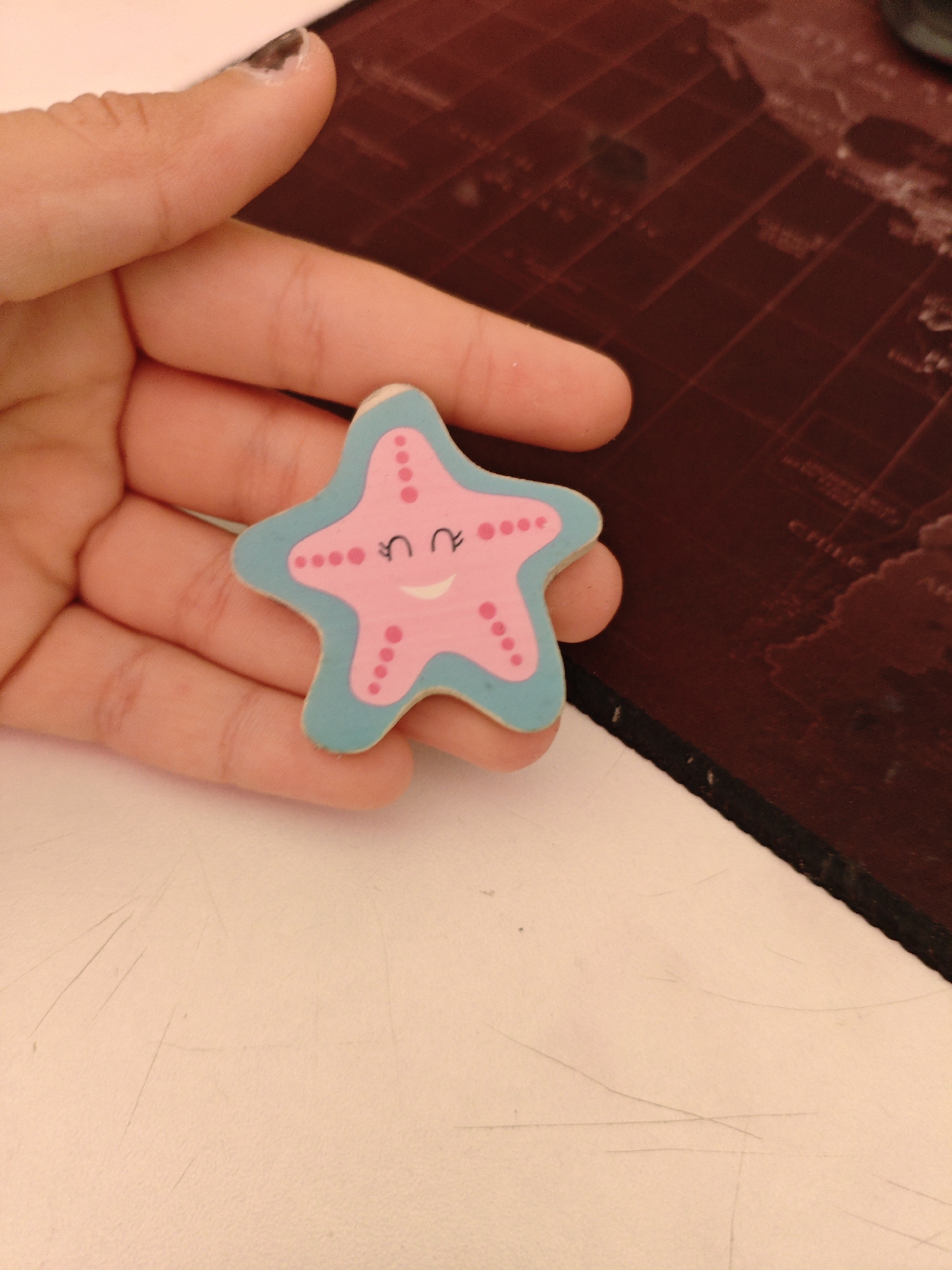 An overhead shot shows a person's left hand holding a pink and blue wooden starfish figurine. The starfish has a smiley face with closed eyes and a smiling mouth. The starfish is positioned on a white surface, which is slightly scratched. Behind the white surface is a dark reddish-brown surface with a map-like pattern. The background surface appears to be a desk or table. The image is sharp and well-lit, with the starfish being the primary subject. The image does not provide any information about the location in the city of Unknown, Unknown.