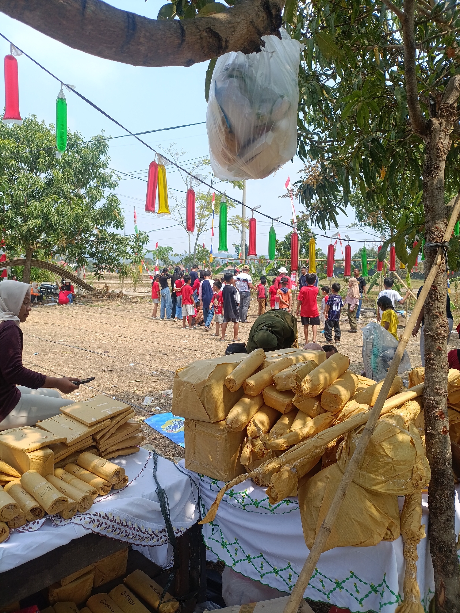 In Cariu, Indonesia, an outdoor scene unfolds under a bright, sunny sky with scattered clouds. The foreground is dominated by stacks of goods wrapped in light brown paper, piled high on tables covered with white cloths adorned with floral embroidery. A woman wearing a hijab and a maroon shirt is seated in the lower left, holding a mobile phone. Behind her, a diverse group of people, including children and adults, are gathered in a dusty, open area. Many of the children are wearing red shirts. Decorative elements, such as elongated, colorful balloons in red, yellow, and green, hang from strings stretched across the area, adding a festive touch. Trees with green foliage provide shade in various parts of the scene. The background reveals a rural landscape with distant hills and what appears to be agricultural land, suggesting the event is taking place outside of a dense urban environment.