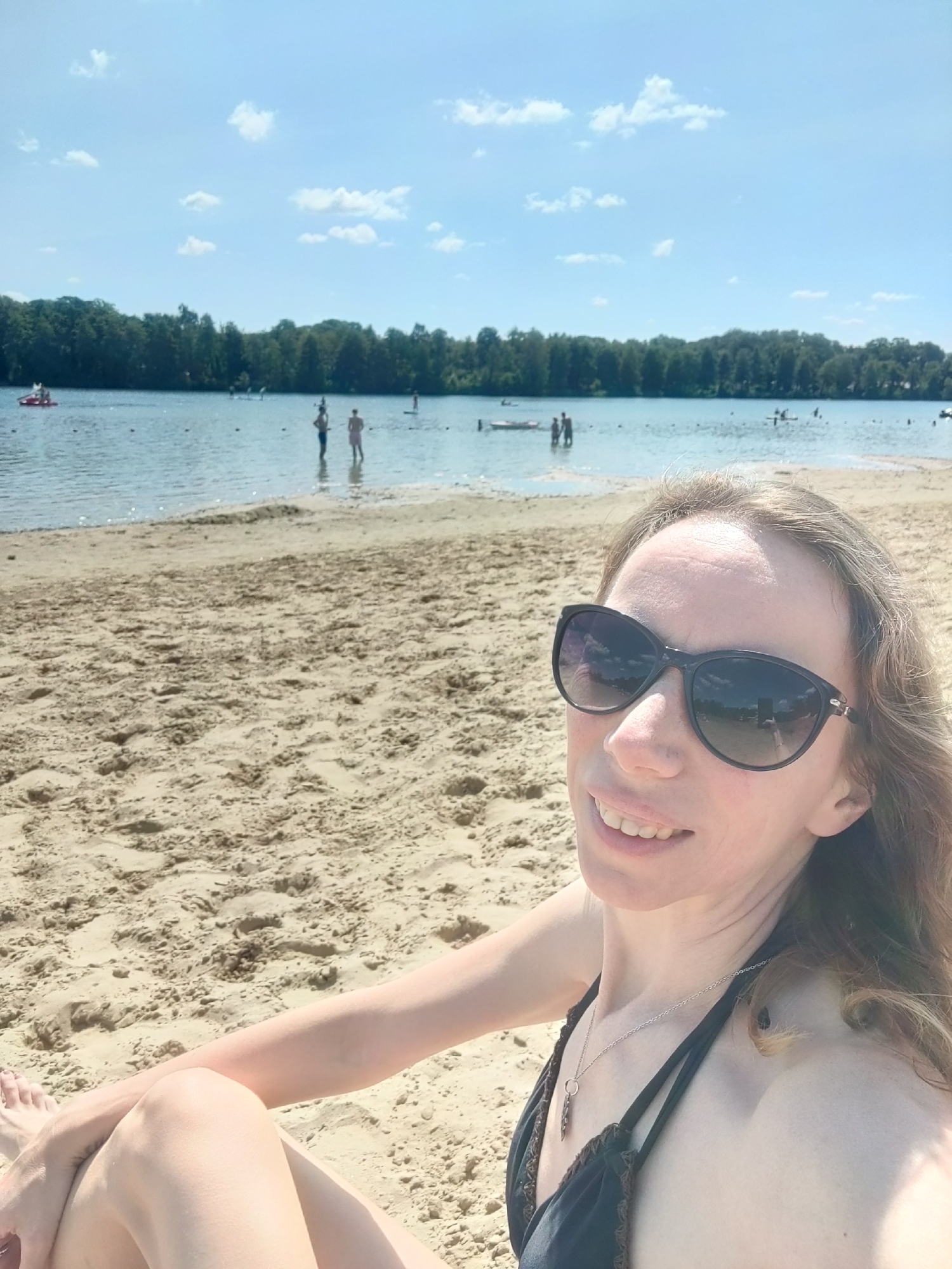 A woman with light brown hair and sunglasses is sitting on a sandy beach, with a lake and trees in the background. The lake is populated with several people, some wading in the water and others on paddle boats. The sky is blue with scattered white clouds. The overall scene is one of a summer day at a recreational lake.