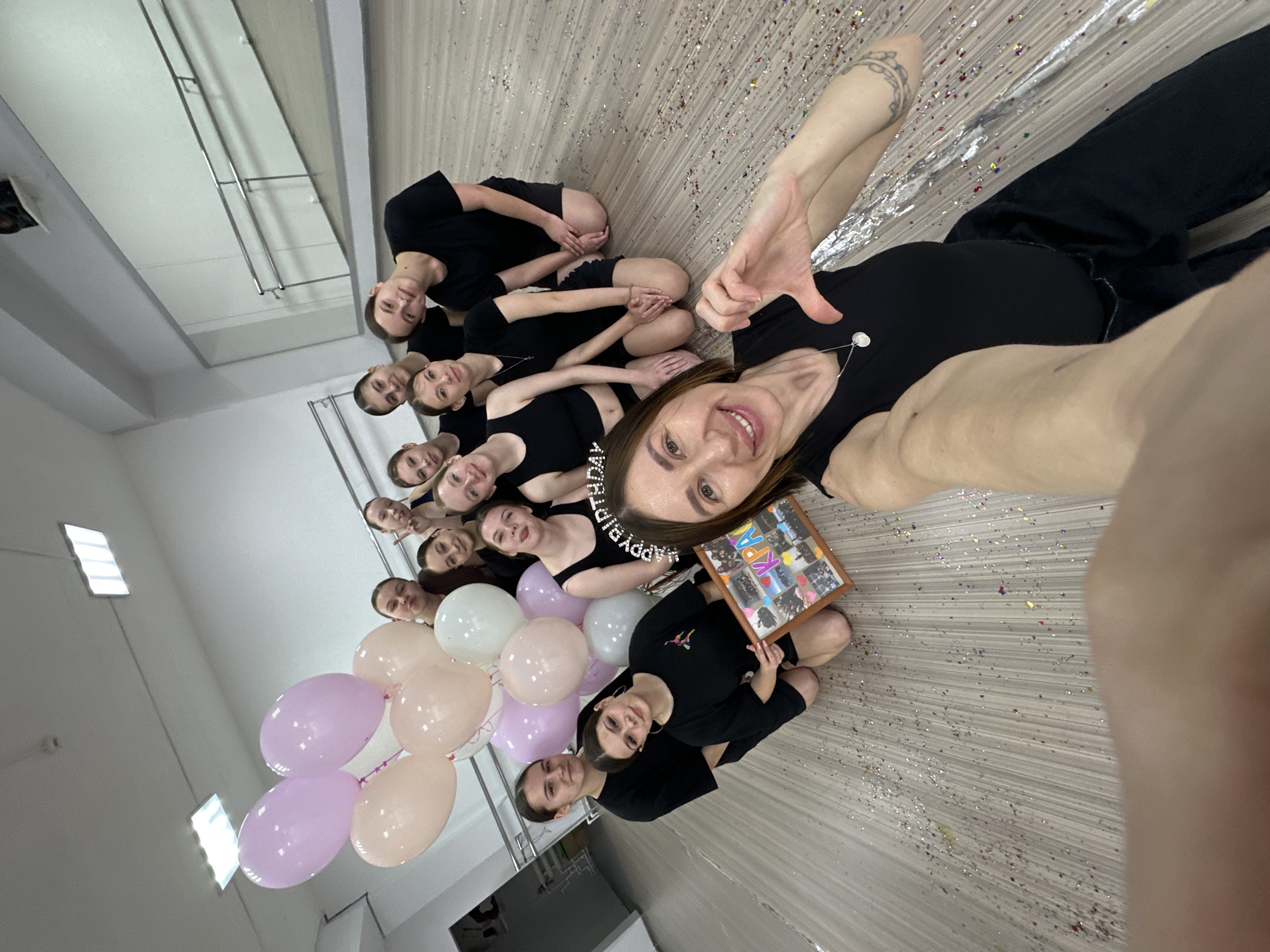 A group of thirteen young women are posing for a selfie in a room scattered with confetti. All are wearing black clothing. The woman taking the selfie has a "Happy Birthday" headband and is holding a framed collage. Pink and white balloons are near the group. The location is in Votkinsk, Russia, discernible from the letters "KPA" visible on the collage, an abbreviation referring to a dance group in the city. A mirror, windows, and tiled floors are visible in the room.