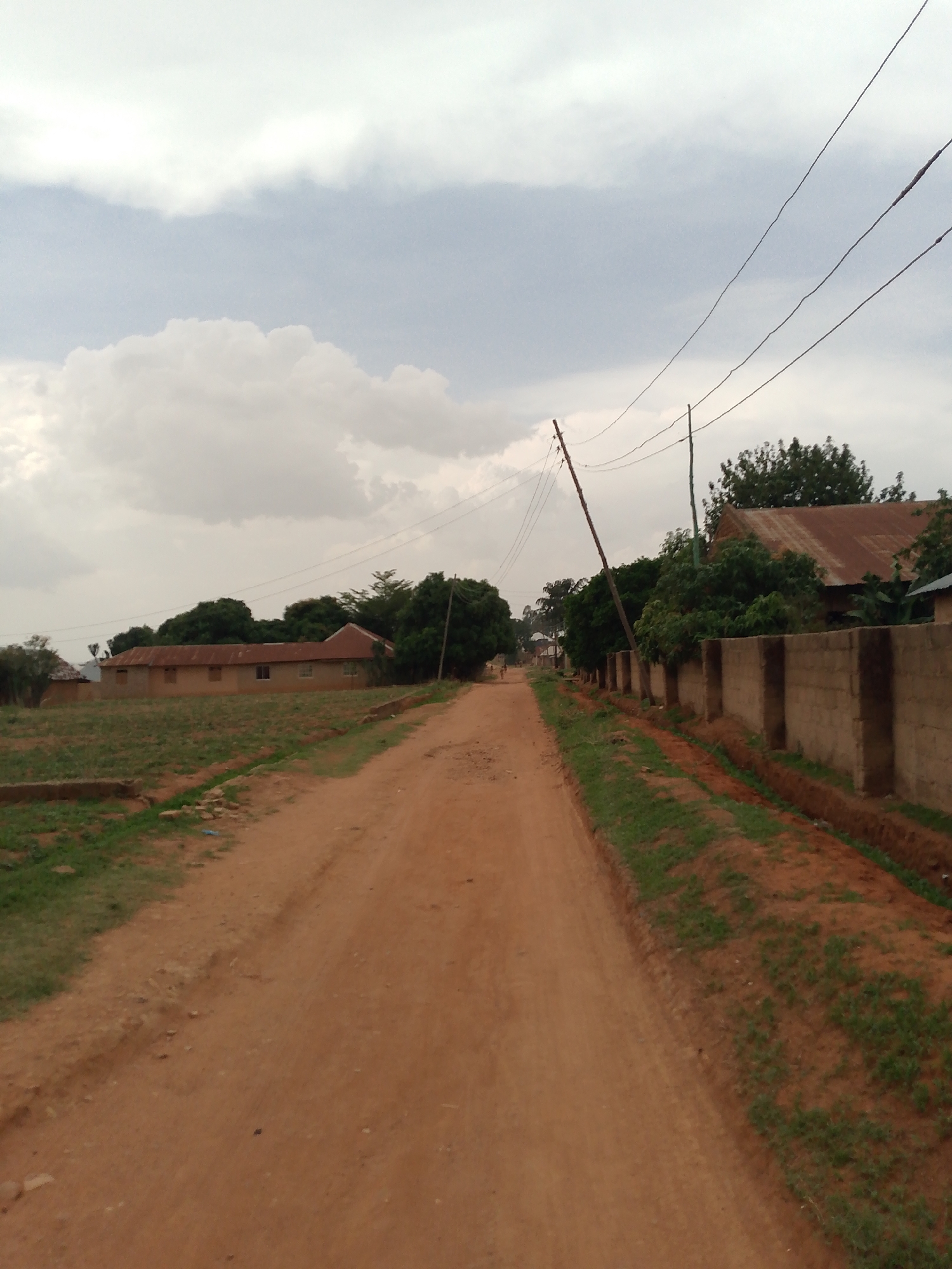 The image shows a dirt road in what appears to be a rural or developing area of Jos, Nigeria. The road is unpaved and reddish-brown in color. On the left side of the road is a field with sparse vegetation. A building with a red roof is visible in the distance. On the right side of the road is a concrete block wall, and a ditch running along the wall. Power lines stretch across the sky. The sky is overcast with gray clouds.